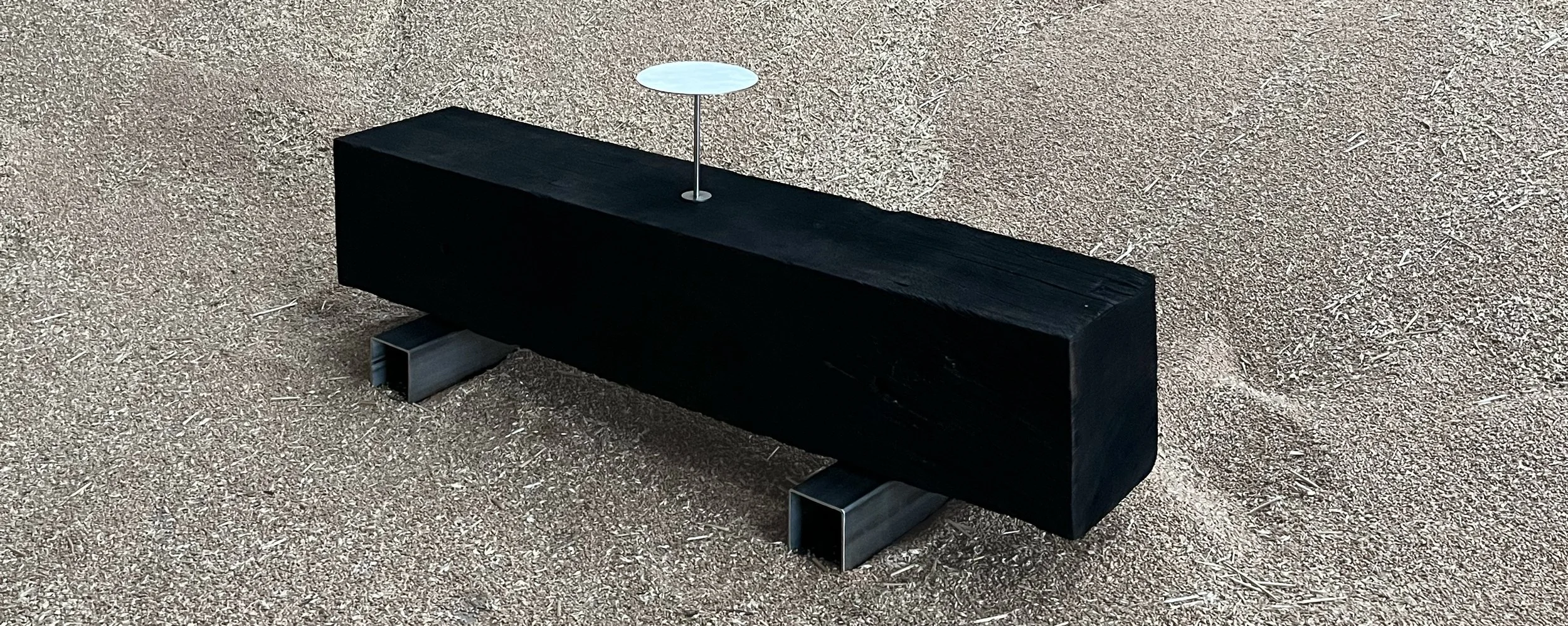 Modern outdoor bench on gravel with a small, thin, circular, white lamp attached to it.
