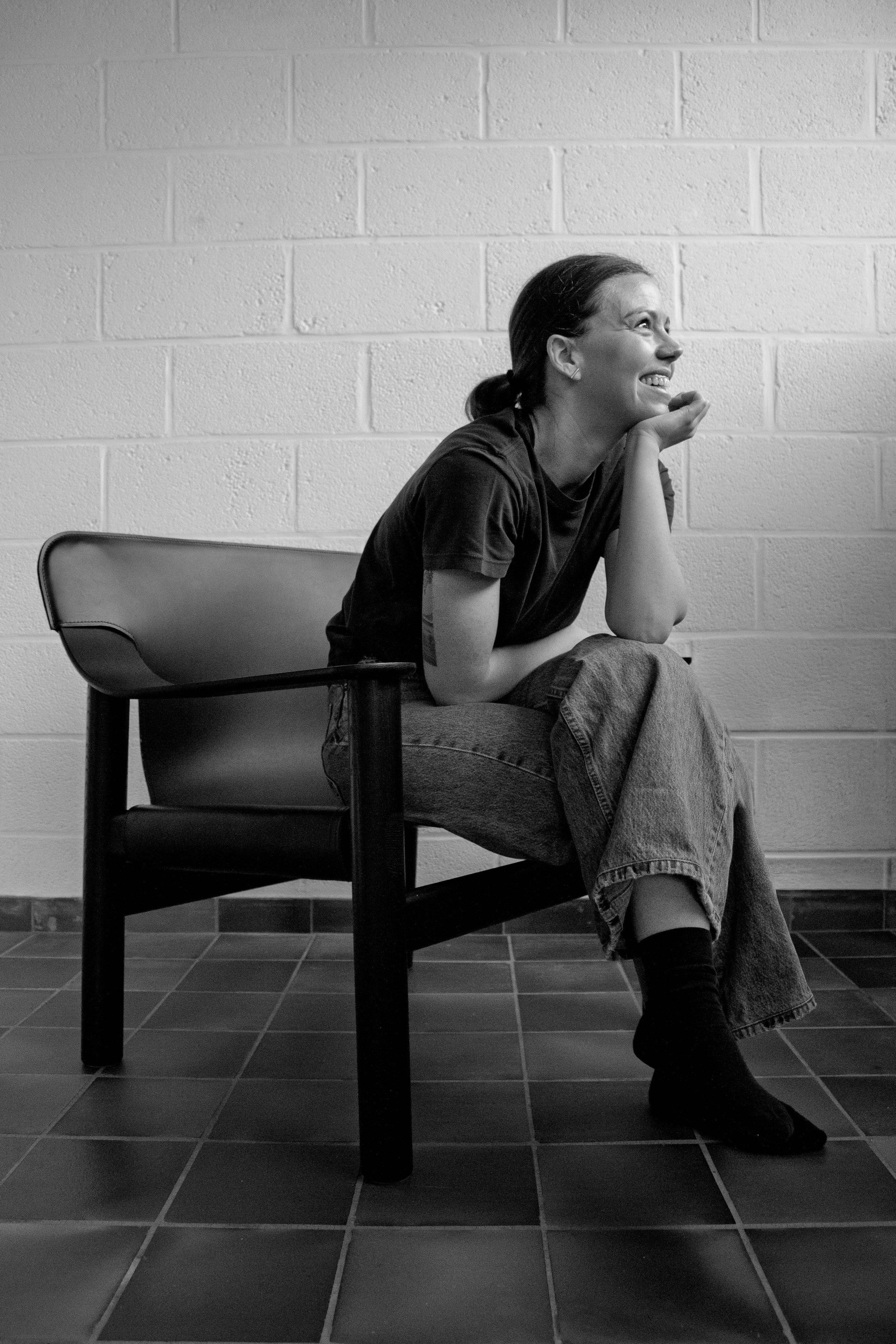 A woman sitting on a bench with her legs crossed, smiling, and looking to her right, in front of a white brick wall.