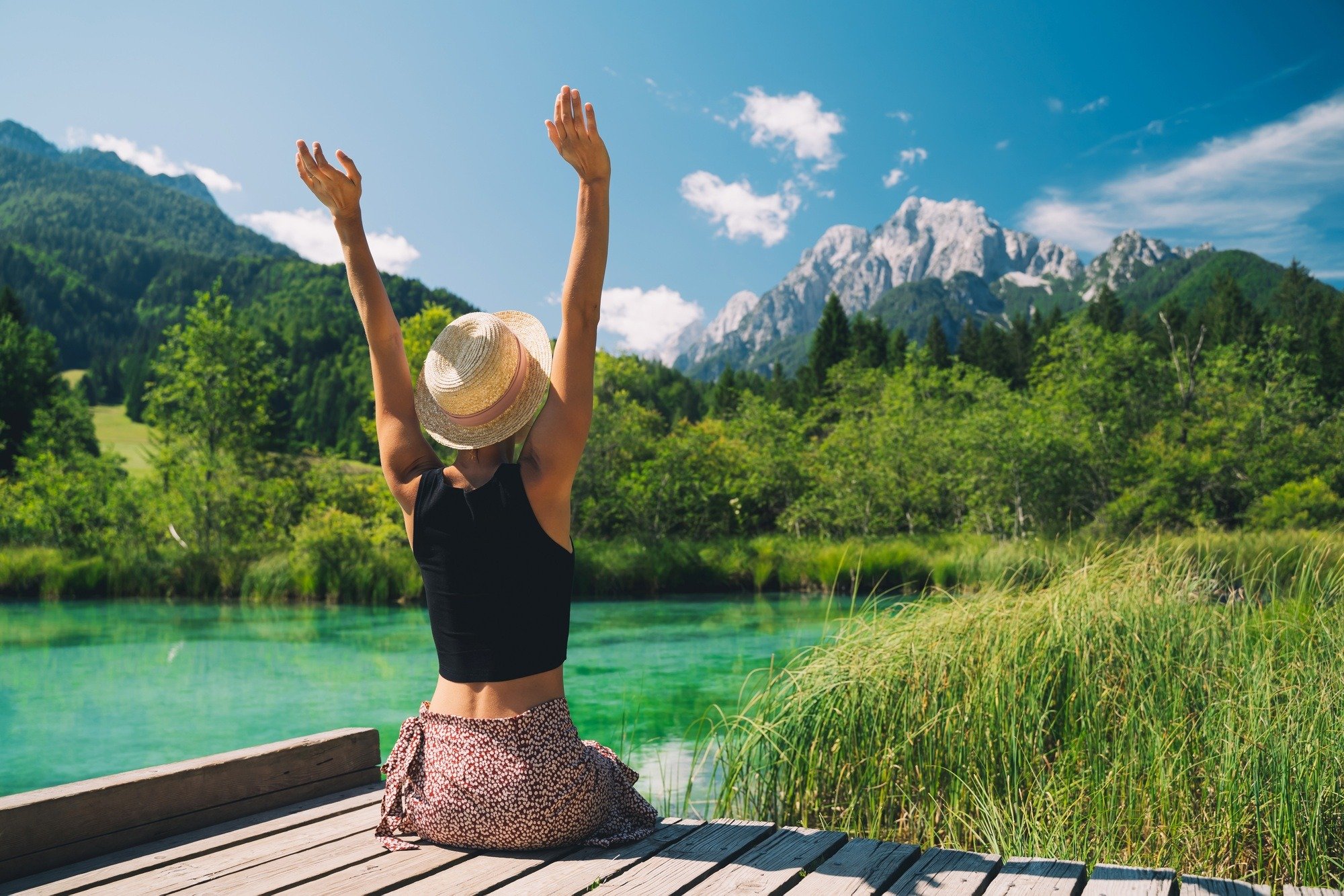 A woman sitting on a wooden dock with her arms raised, facing a mountain landscape with green trees, a lake, and mountains in the background.