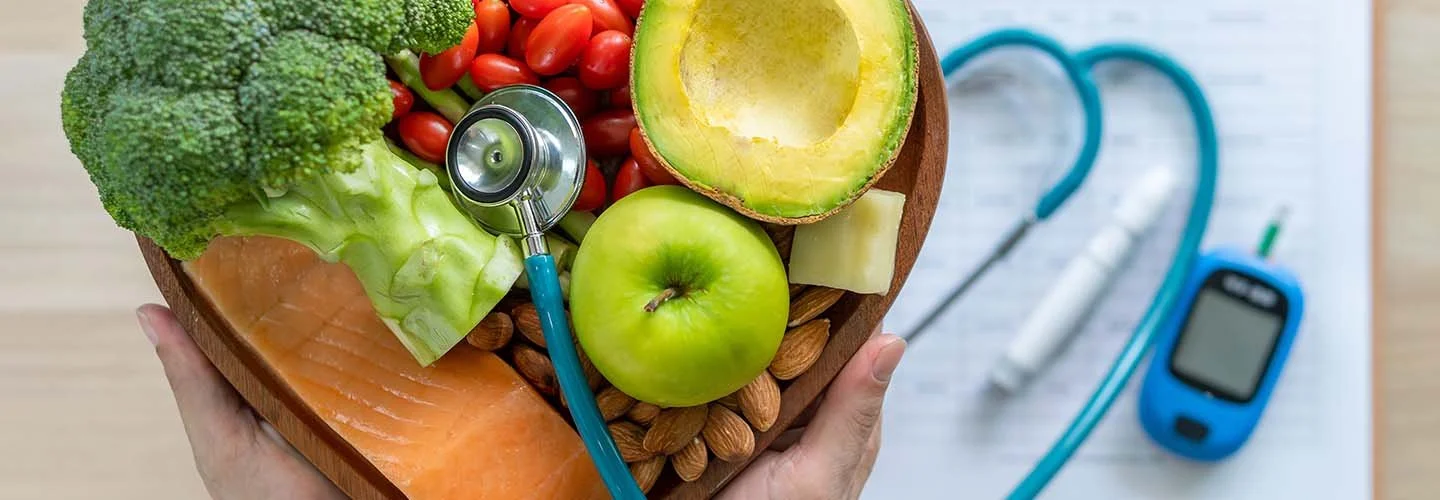 Fresh vegetables, including broccoli, cherry tomatoes, avocado, apple, lettuce, and salmon, arranged in a wooden heart-shaped dish with a stethoscope on top; background shows a glucometer and medical supplies.