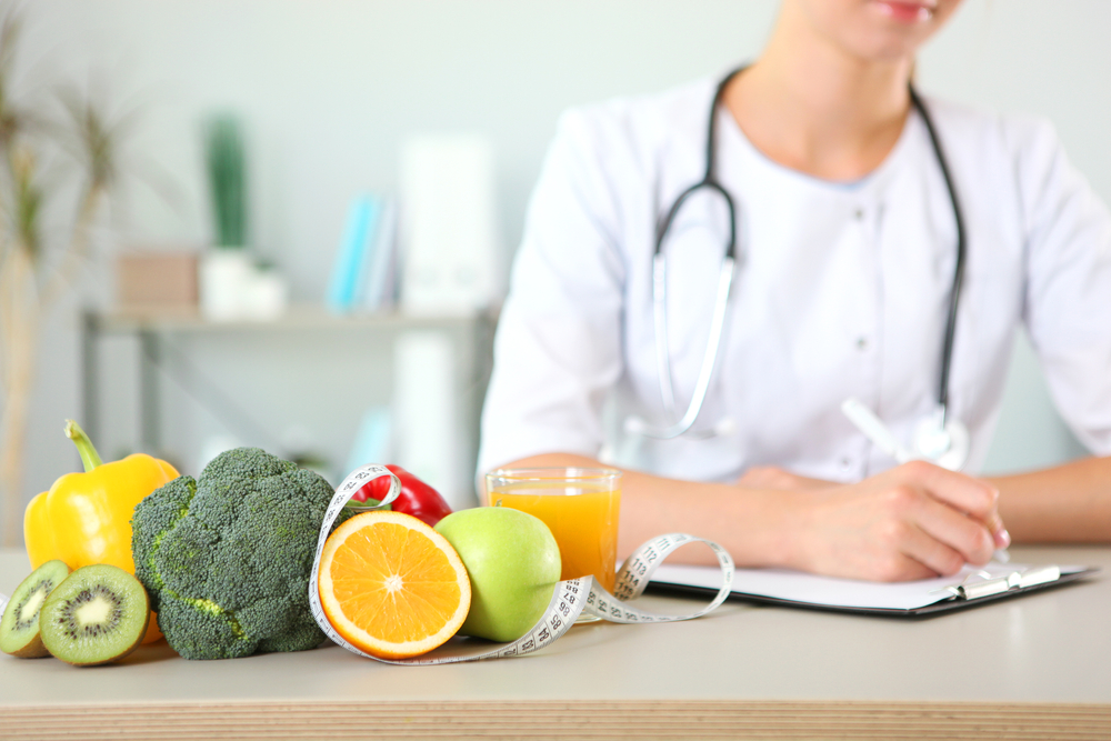 Doctor with stethoscope at a desk with fruits, vegetable, orange juice, and a measuring tape.