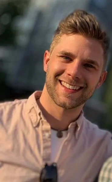 A young man in Fairfax, virginia with short brown hair and a beard smiling outdoors in front of a building, wearing a light-colored button-up shirt.