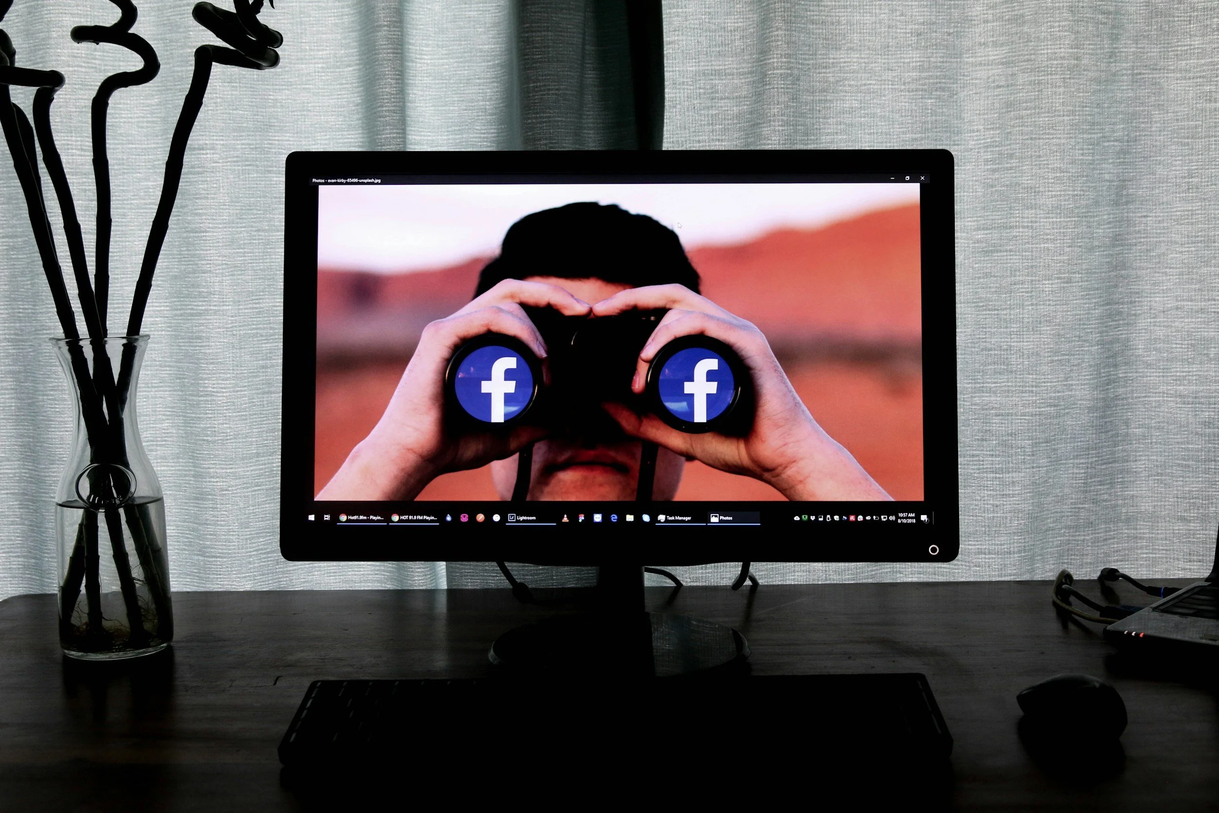 Computer monitor displaying a person holding binoculars with Facebook logos over the lenses, on a dark wooden desk with a glass vase containing black sticks on the left and electronic devices on the right.