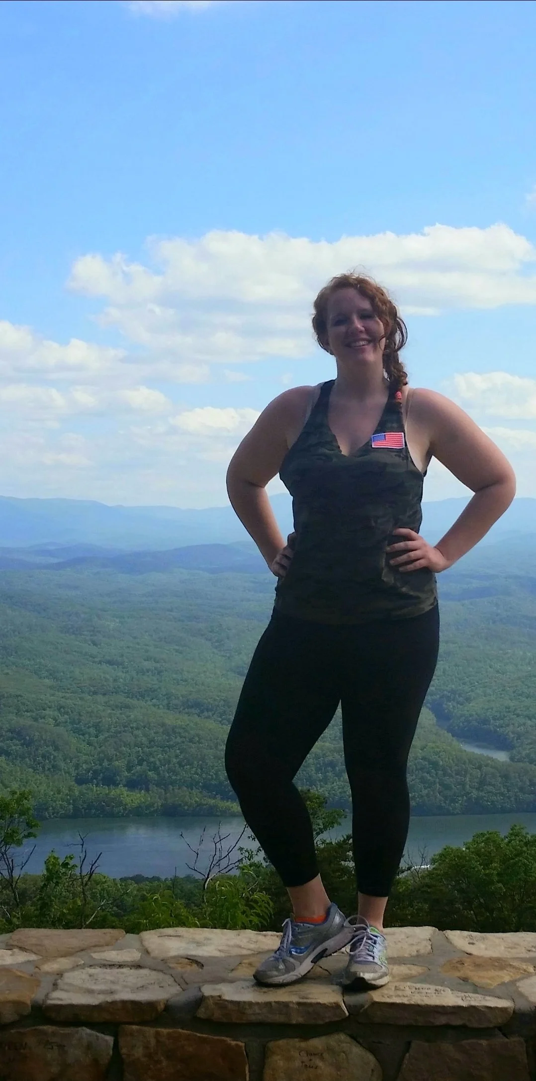 A woman with curly red hair standing on a stone ledge outdoors on a mountain, with a view of green forests, a river, and a blue sky with clouds in the background.