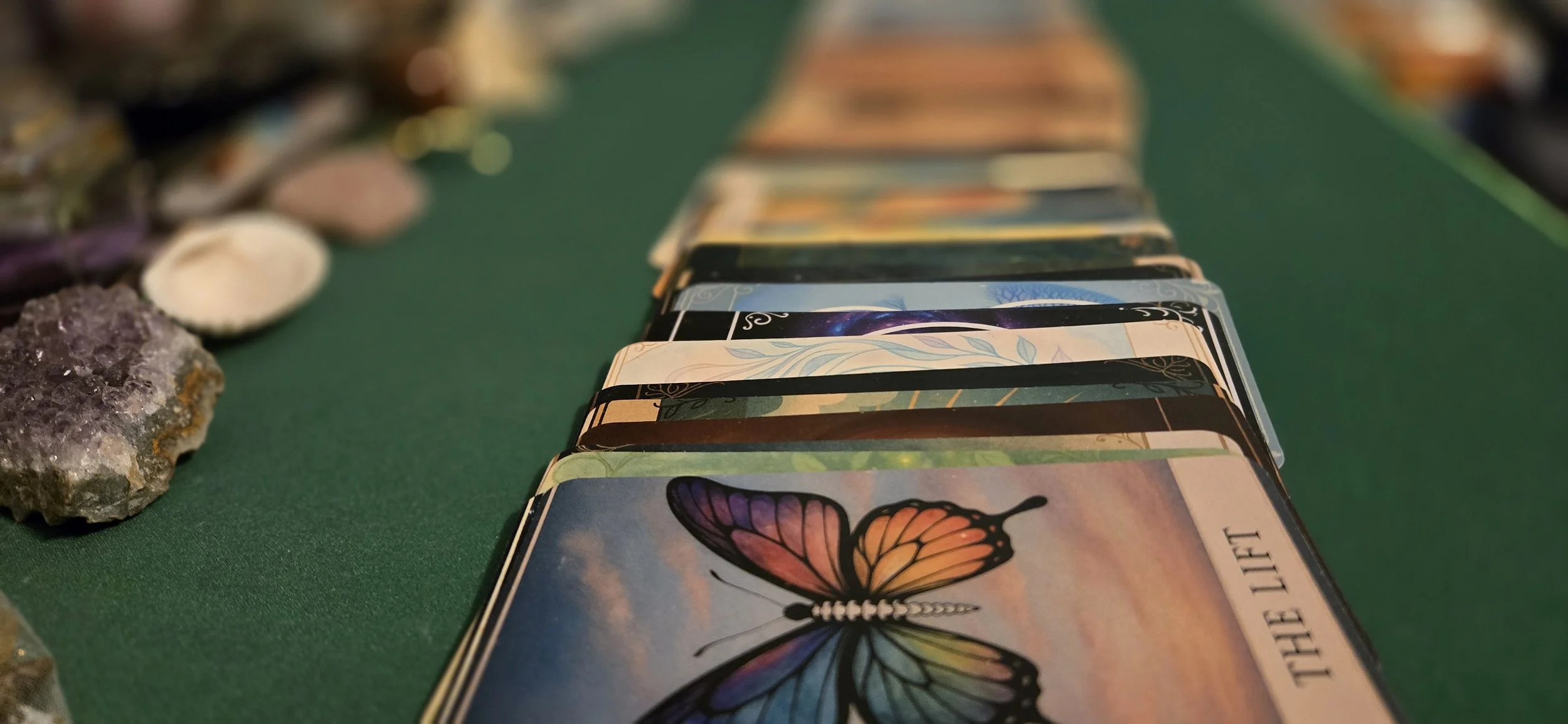 A spread of tarot cards placed on a green cloth, with crystals and shells nearby, featuring a card with a colorful butterfly illustration and the partially visible text "THE LIFT".