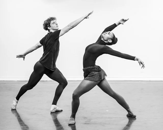 Two ballet dancers practicing in a studio, performing dramatic poses with arms extended and legs in wide stances.