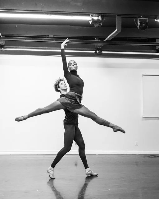 Ballet dancers practicing in a dance studio with one dancer lifting another in an arabesque pose.