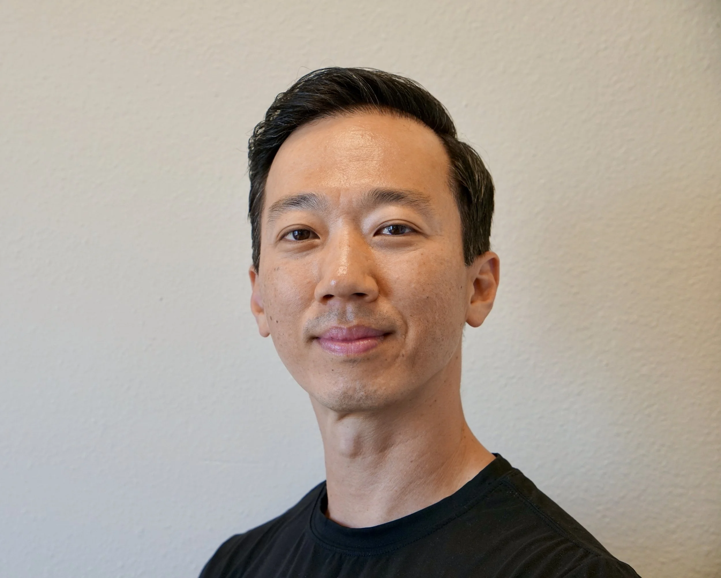 Close-up of a young man with short black hair, looking at the camera, wearing a black shirt, against a plain light-colored background.