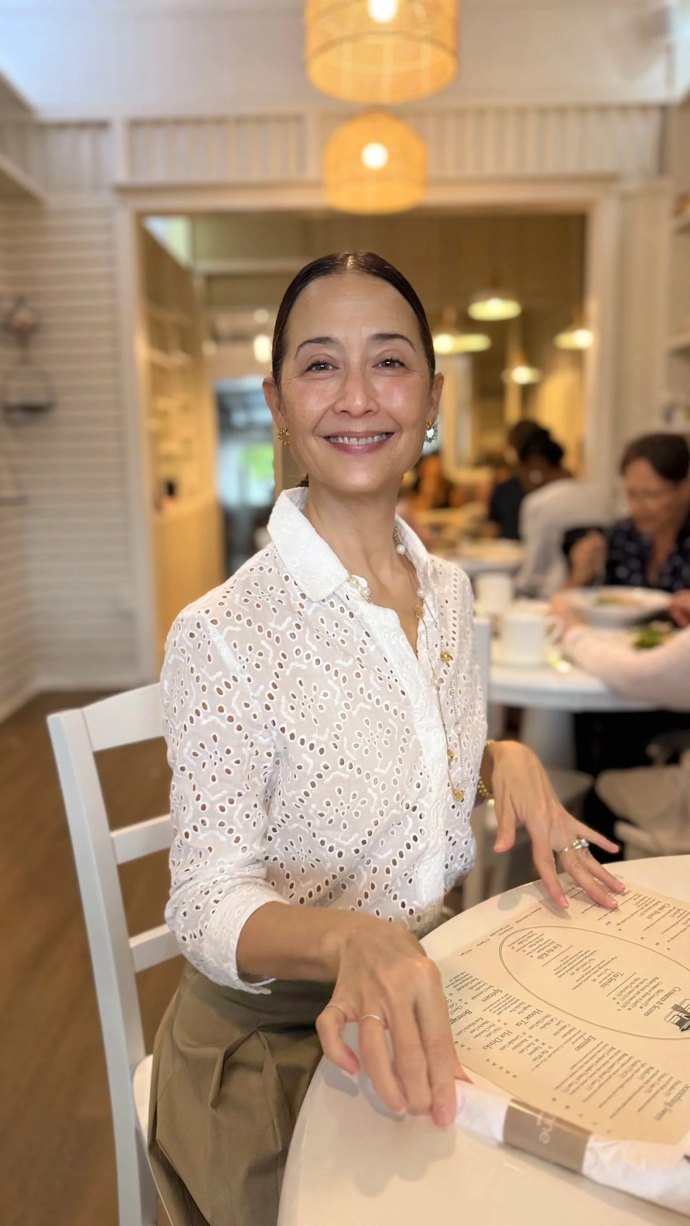 A woman with dark hair pulled back, smiling, sitting at a table in a bright restaurant, wearing a white eyelet blouse and jewelry, with a menu in front of her, and people in the background dining.
