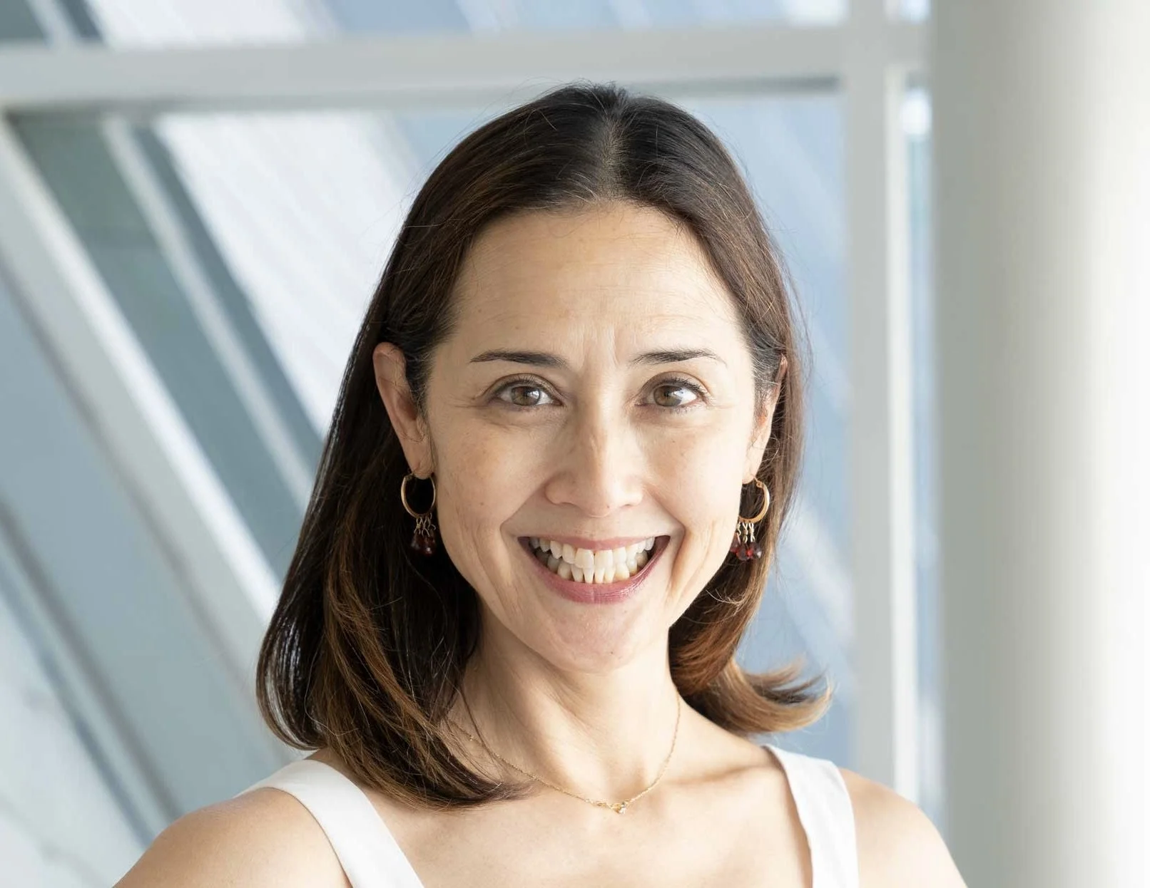 Close-up photo of a woman with shoulder-length brown hair, wearing gold earrings and a white sleeveless top, smiling warmly in front of a modern architectural background with glass and metal.