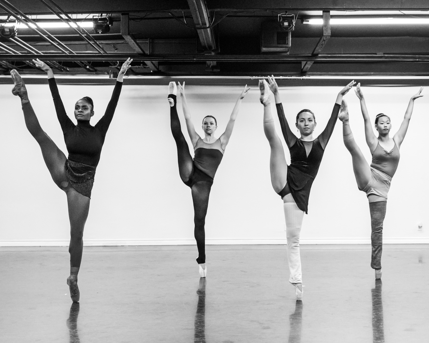 Four female ballet dancers practicing ballet poses in a studio. They are standing on one leg with the other leg extended high, and arms raised in various positions. The photo is in black and white.
