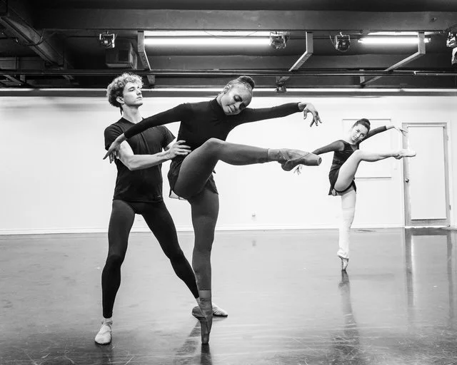 Three ballet dancers practicing in a dance studio, with one dancer being supported by another while performing a leg extension and the third dancer stretching.