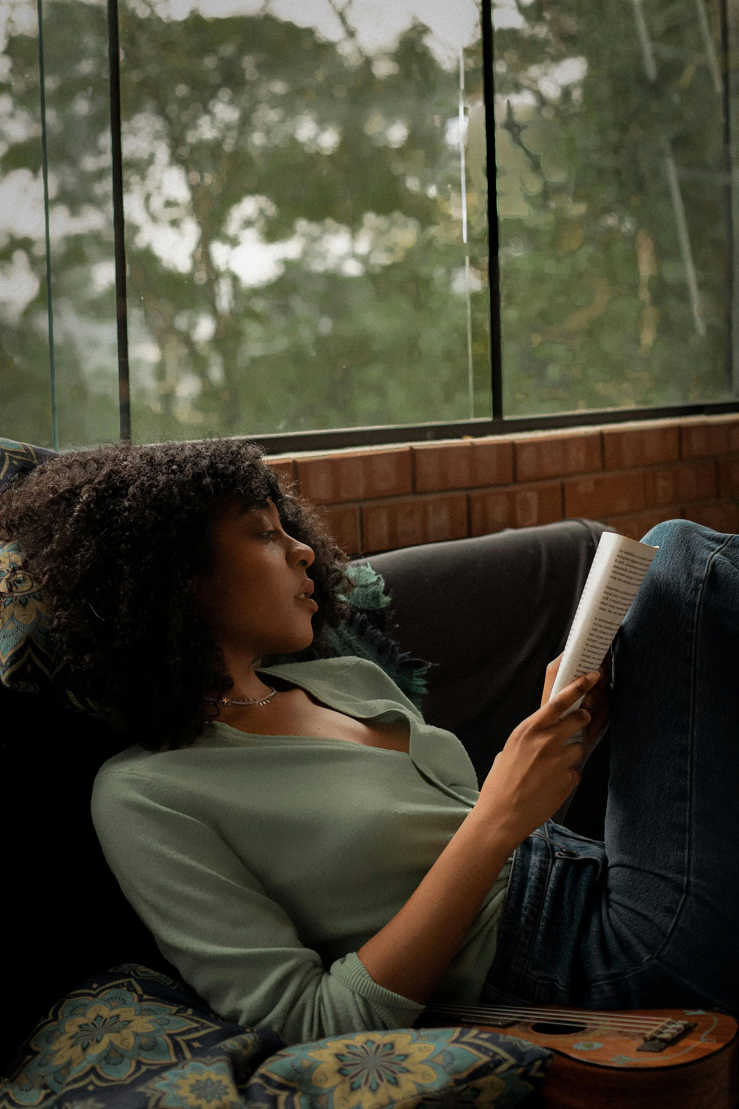 A young woman with curly hair is relaxing on a black couch, reading a book while leaning back, with a window and trees visible outside.