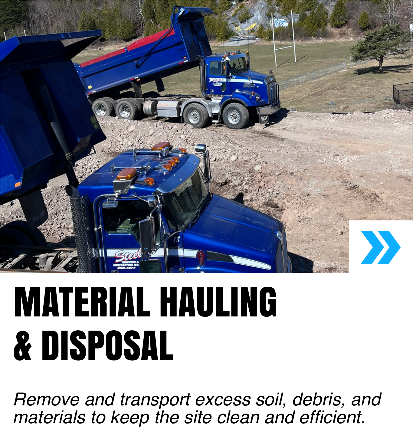 Two large blue and red dump trucks working on a construction site, moving soil and debris. There is a grassy area with trees and a baseball field in the background.