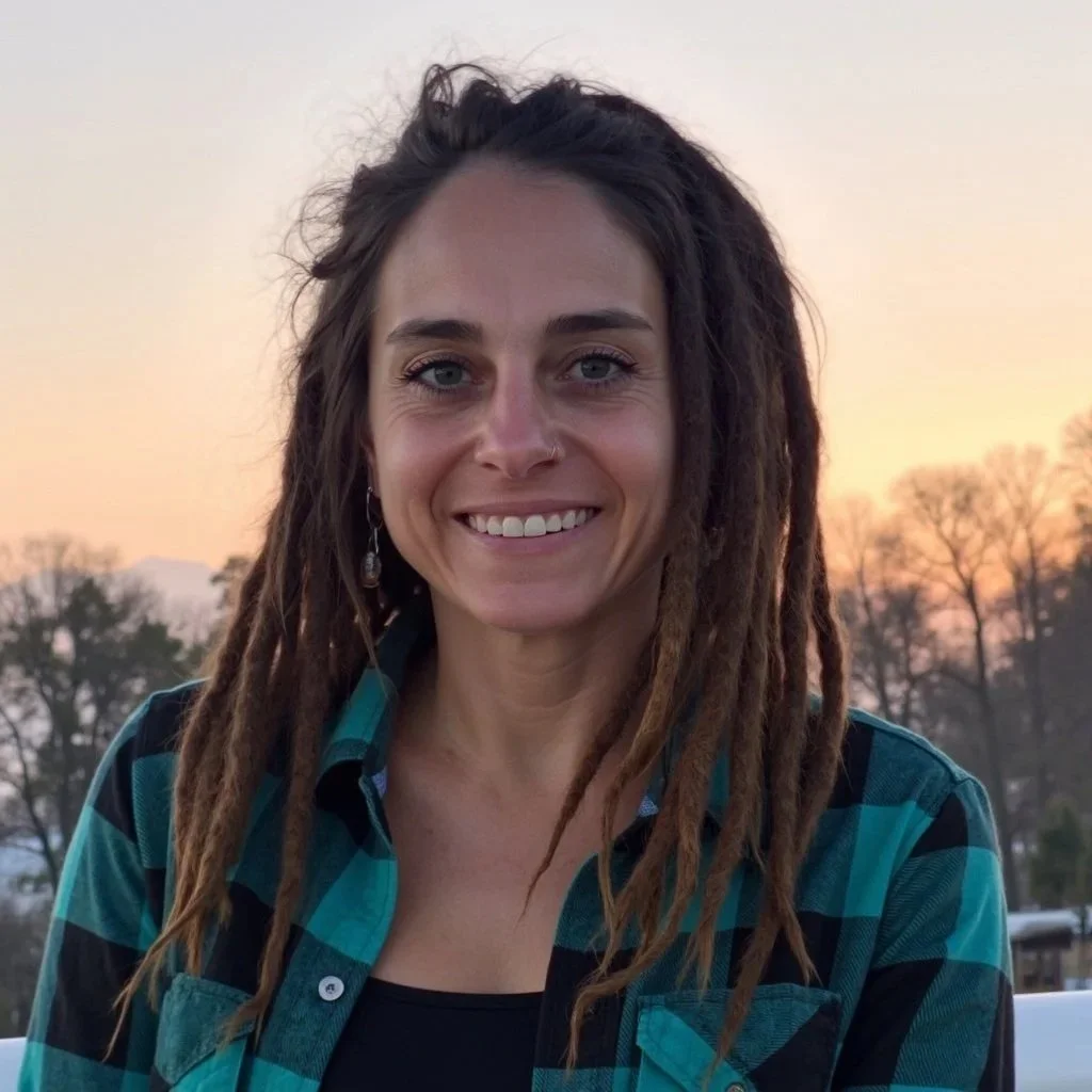 A woman with dreadlocks and a nose piercing, smiling outdoors during sunset, wearing a blue checkered shirt.