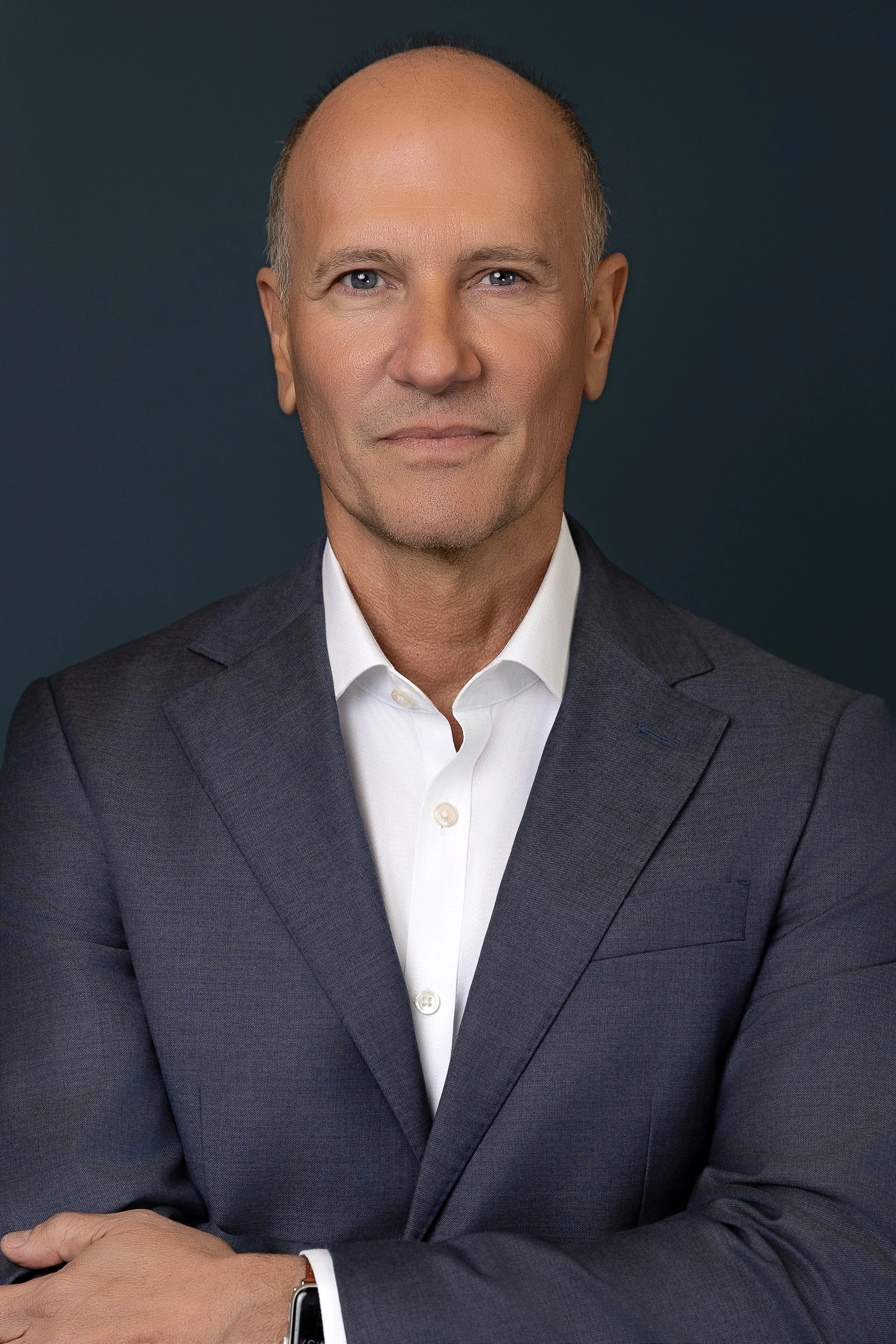 A middle-aged man in a dark blue suit and white shirt looks directly at the camera with a neutral expression. He has a receding hairline and is standing against a dark background.