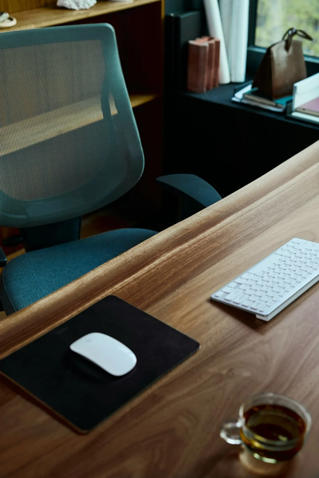 A tidy wooden office desk with a white wireless mouse on a black mouse pad, a white wireless keyboard, a glass cup of coffee, and a teal office chair with a mesh back. In the background, a window, books, and bags are visible.