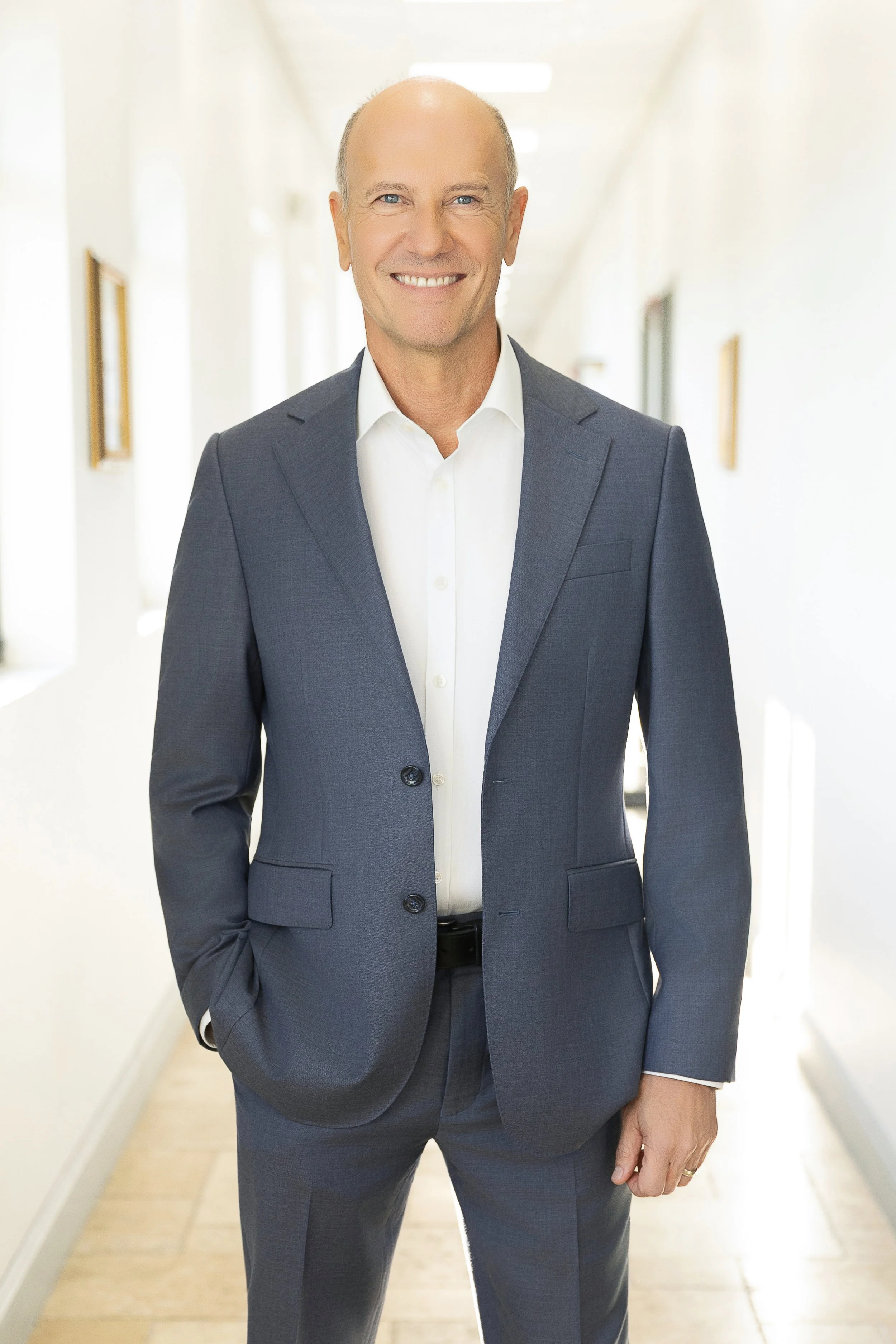 A smiling middle-aged man in a navy suit and white shirt, standing in a bright hallway.