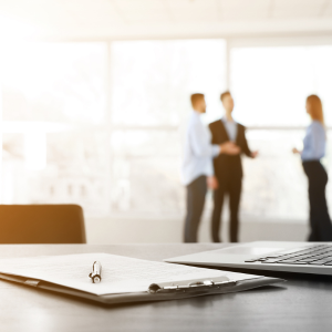 Blurred image of three people in business attire having a conversation in an office with large windows in the background, with a desk, notebook, and laptop in the foreground.