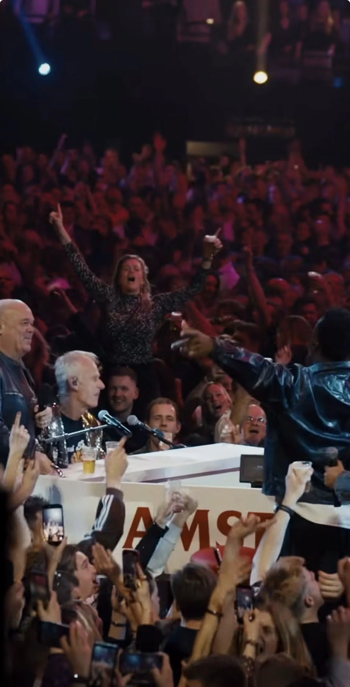 Crowd at a lively concert or event, with a woman raising her arms enthusiastically, many audience members holding up phones, and a man speaking or singing into a microphone on stage.