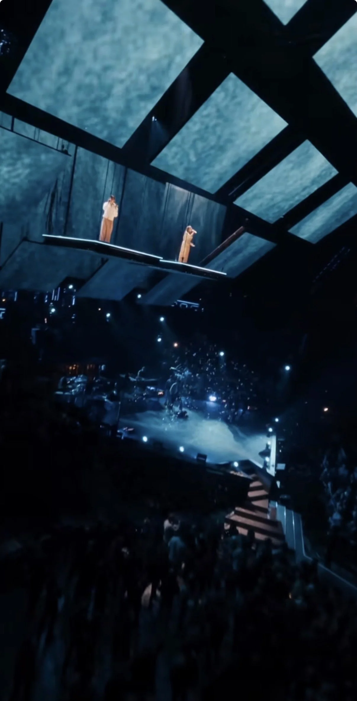 A concert or performance stage with two performers on an elevated platform, and a large crowd of audience members below.