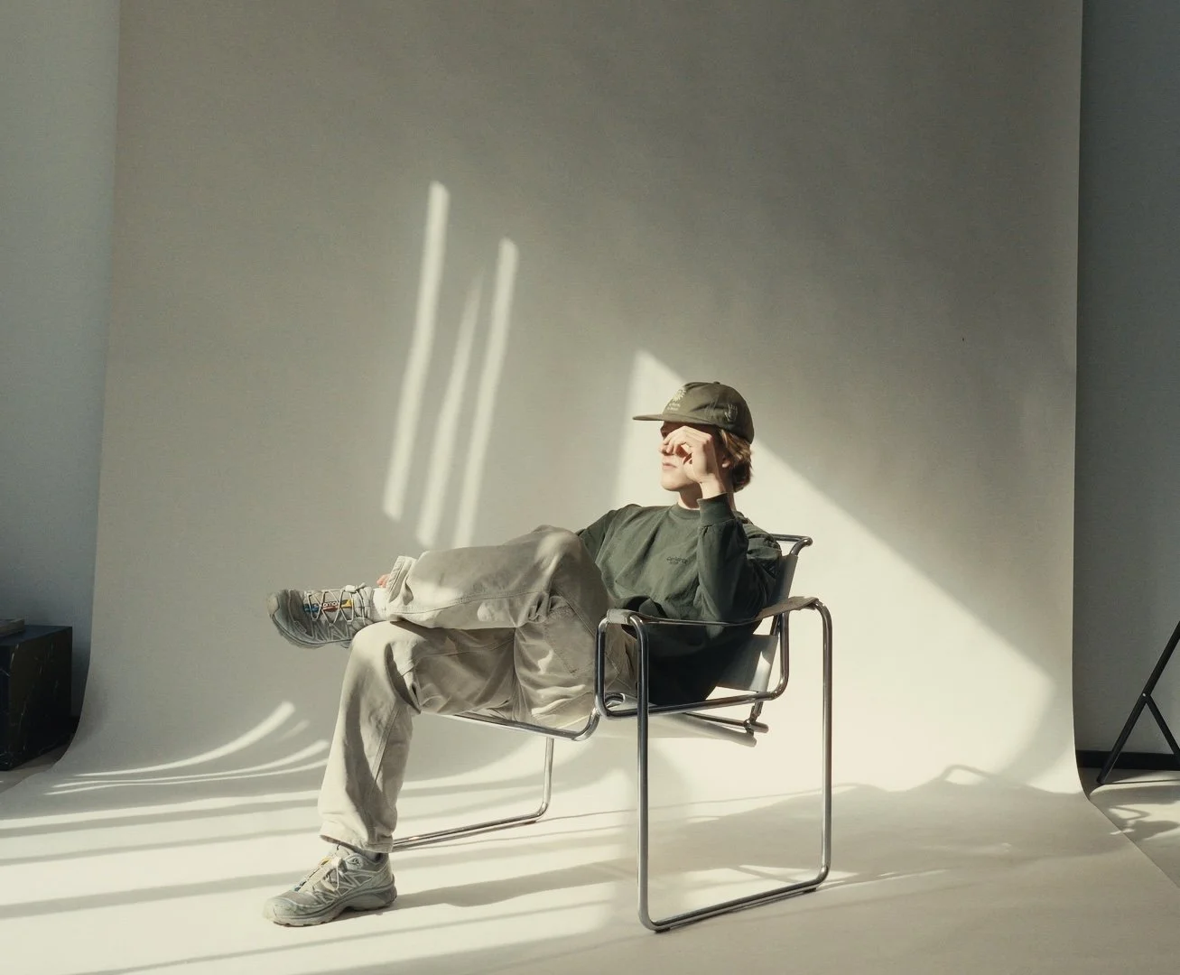 A person with long hair wearing a cap, sunglasses, a dark long-sleeve shirt, and light-colored pants, sitting in a modern armchair in front of a white backdrop. Sunlight creates shadows on the backdrop, casting light and shadow patterns.