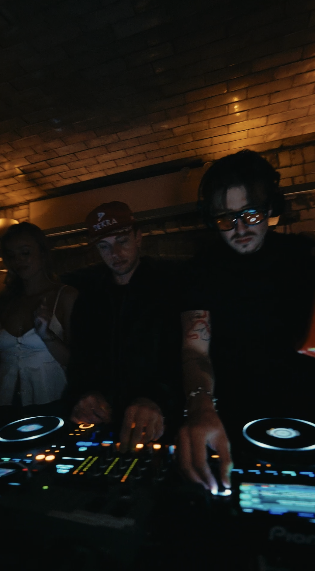 Three people, two men and one woman, standing around a DJ mixer in a dimly lit room with a brick ceiling, part of a nightclub or party setting.