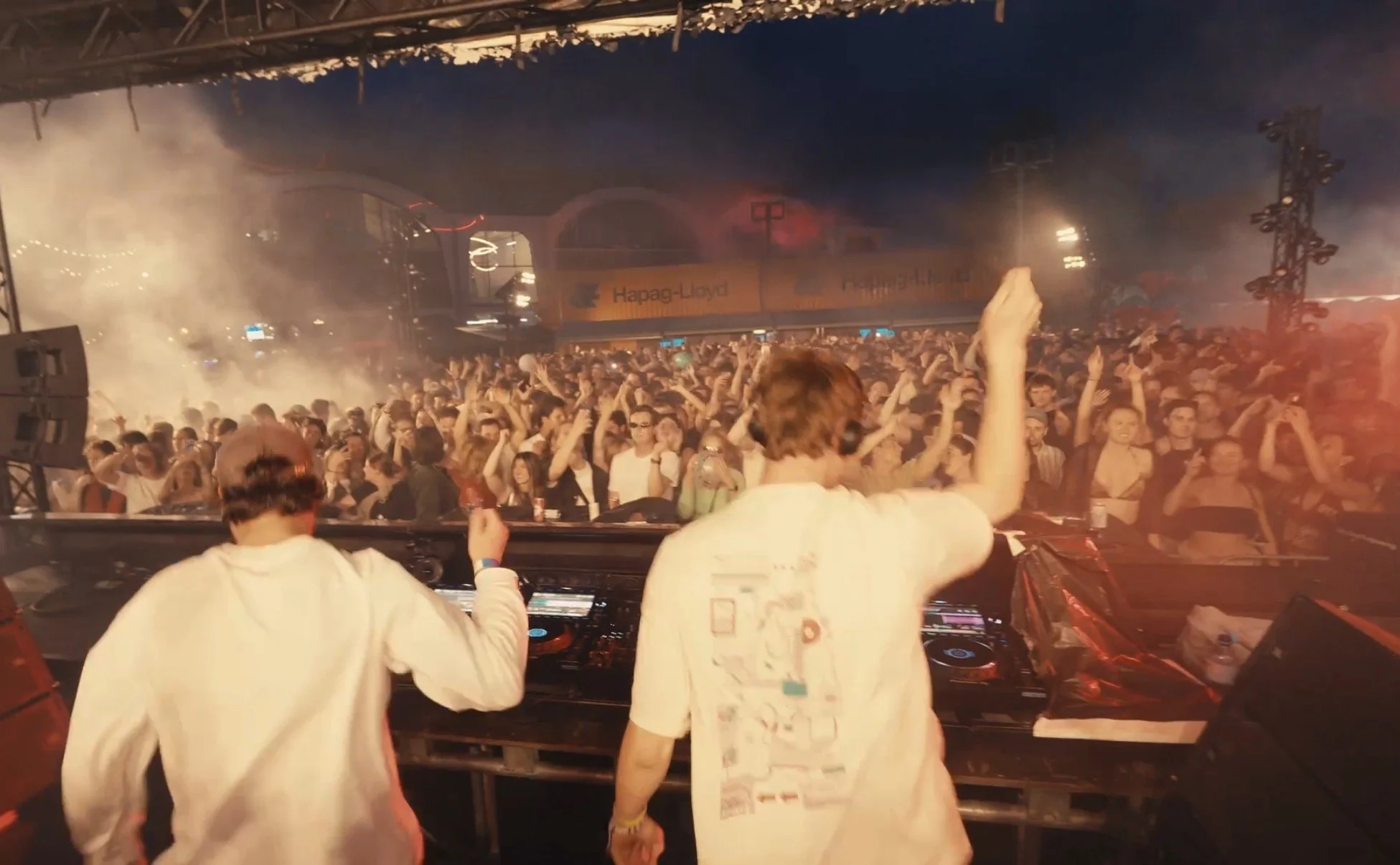 A DJ performing at a crowded music concert with an energetic audience, smoke, and stage lighting.