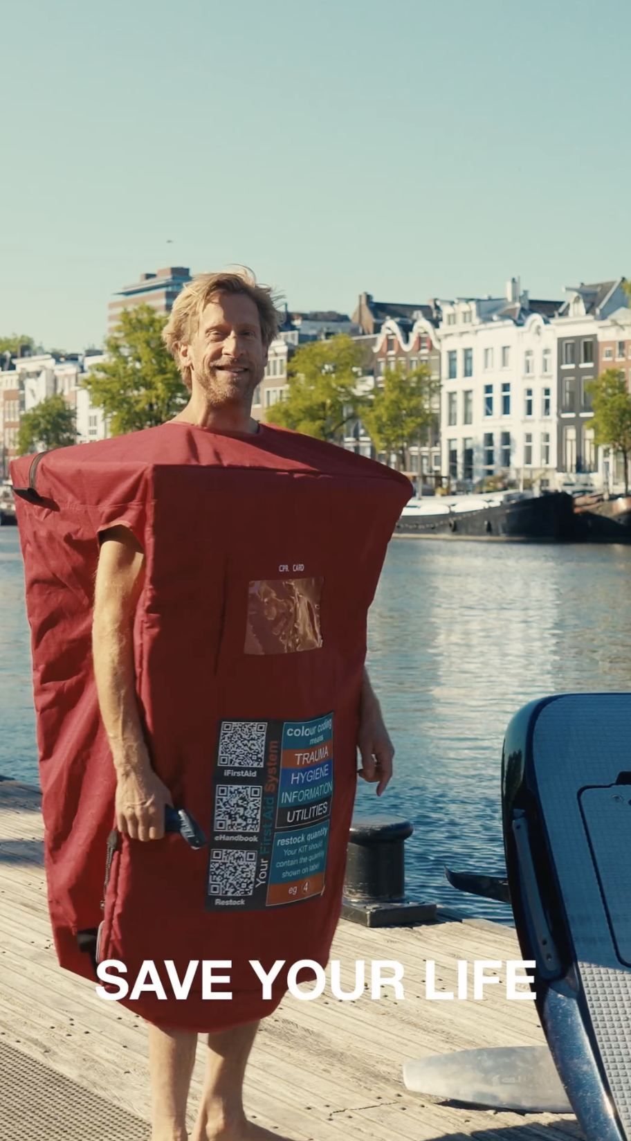 A man wearing a CPR training dummy suit stands on a wooden dock near water, smiling. The suit is red with emergency response information and QR codes. The background features colorful European-style buildings, trees, and boats on the water. Caption reads "SAVE YOUR LIFE."