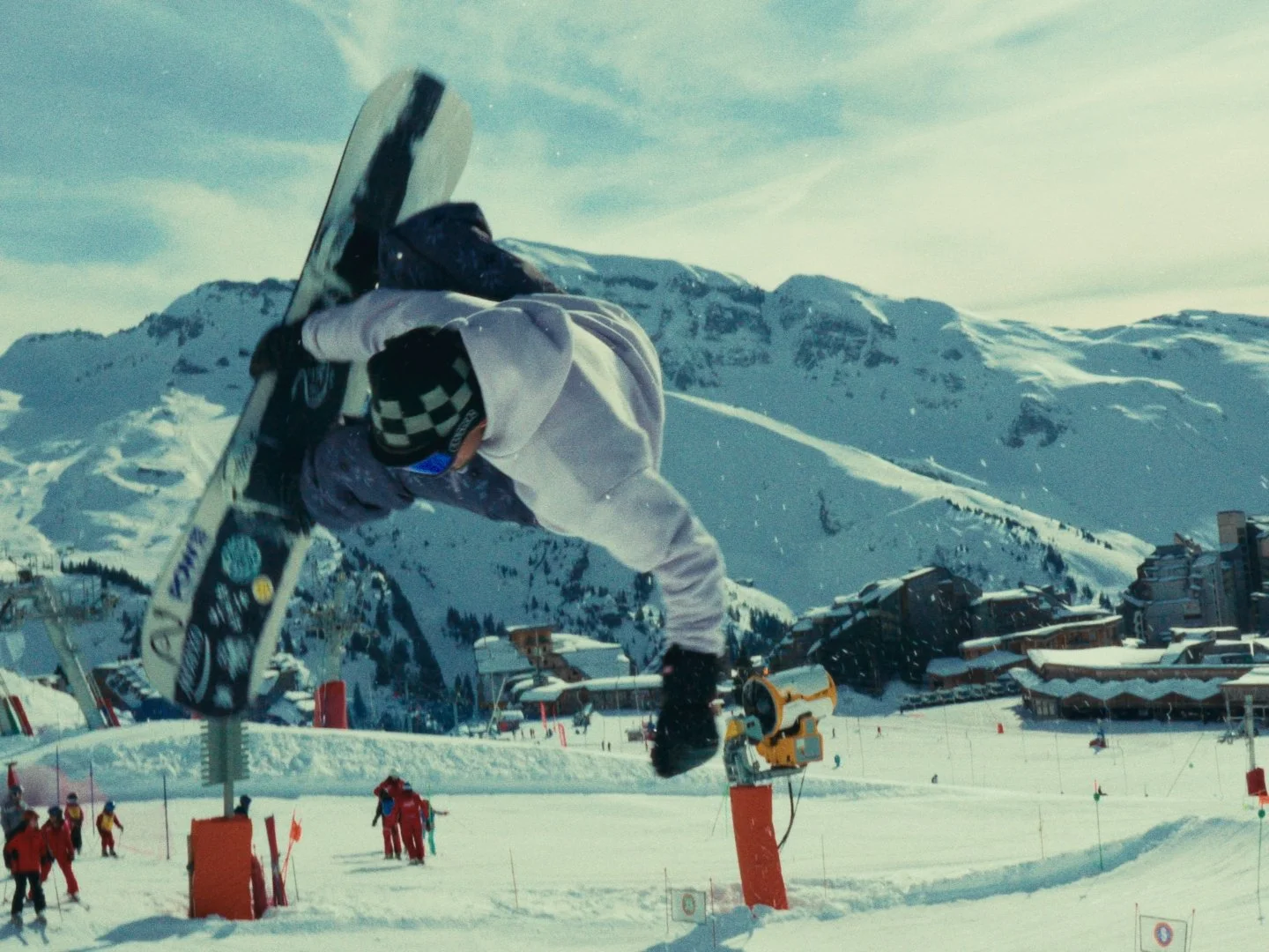 Snowboarder performing a jump in a snowy mountain resort with buildings and ski lifts in the background.