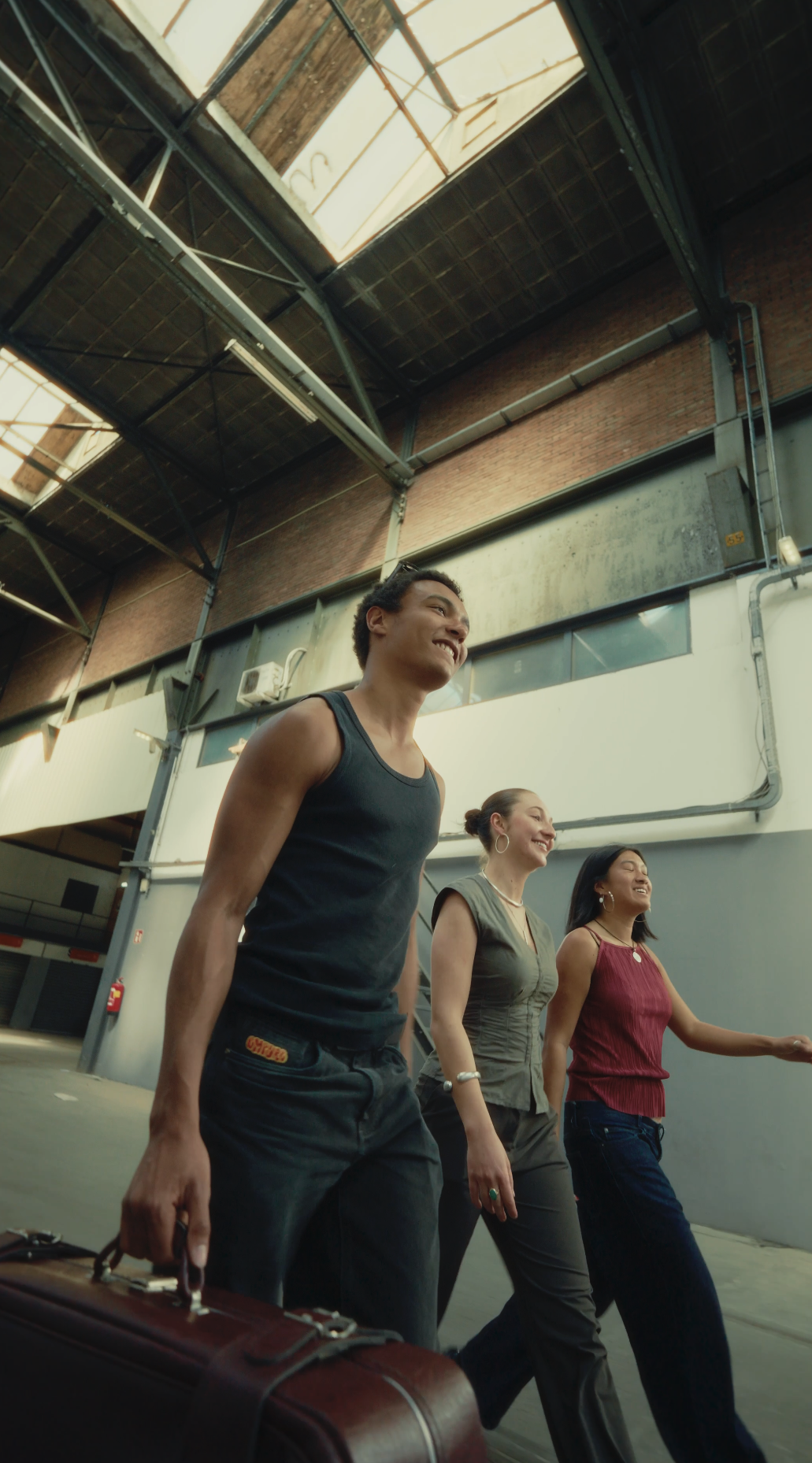 Three diverse people walking together in an industrial-style building with high ceilings and skylights, smiling and carrying a briefcase.