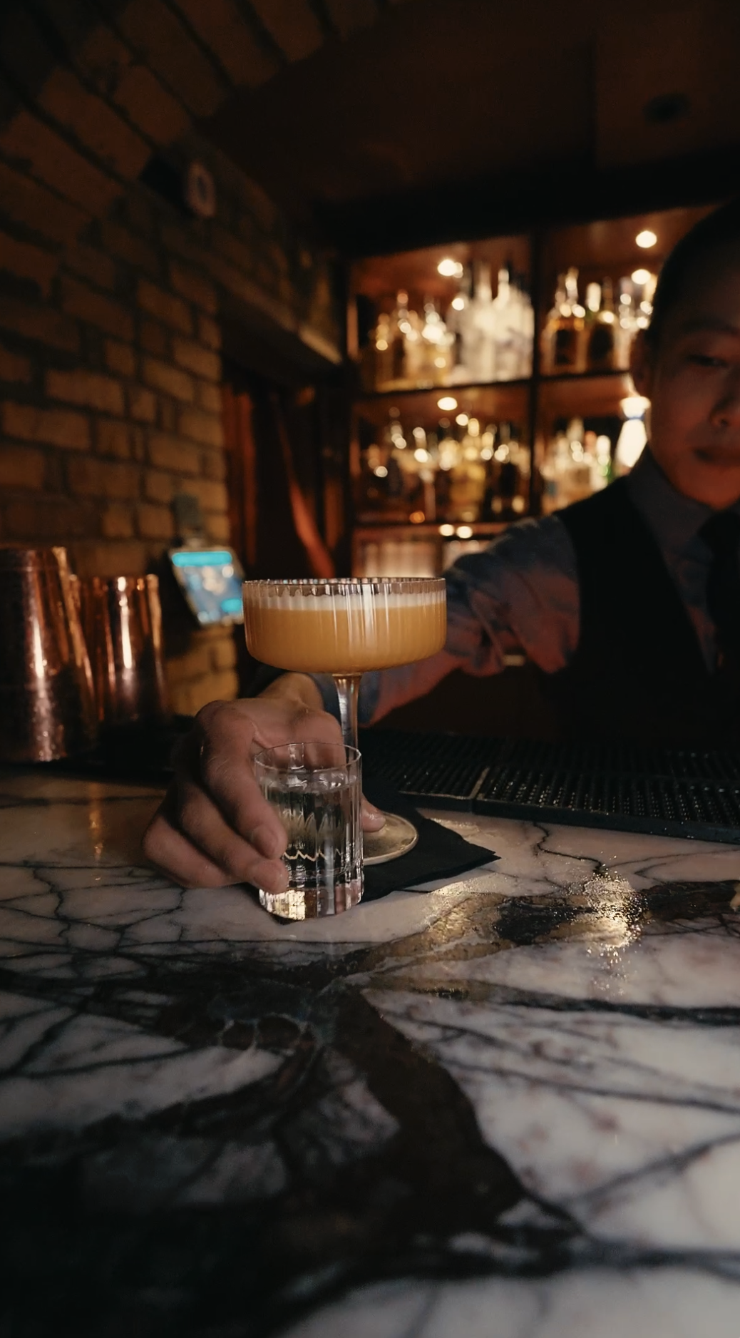 A bartender holding a shot glass in one hand and a cocktail glass with an orange-colored drink in the other, seated at a marble bar counter in a dimly lit bar with brick walls and shelves of liquor bottles in the background.