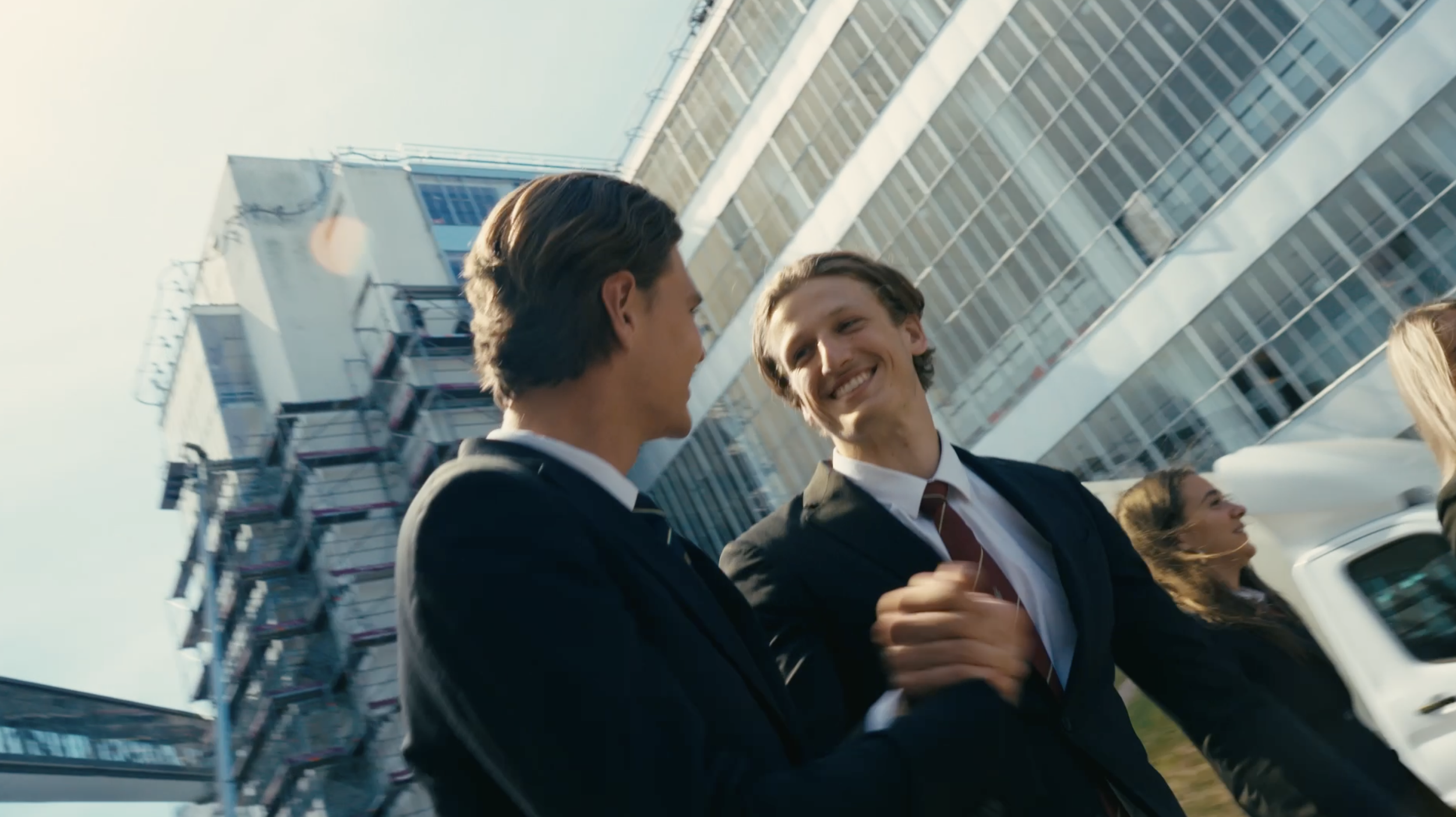 Two young men in suits smiling and talking outside in an urban area with tall modern buildings in the background.