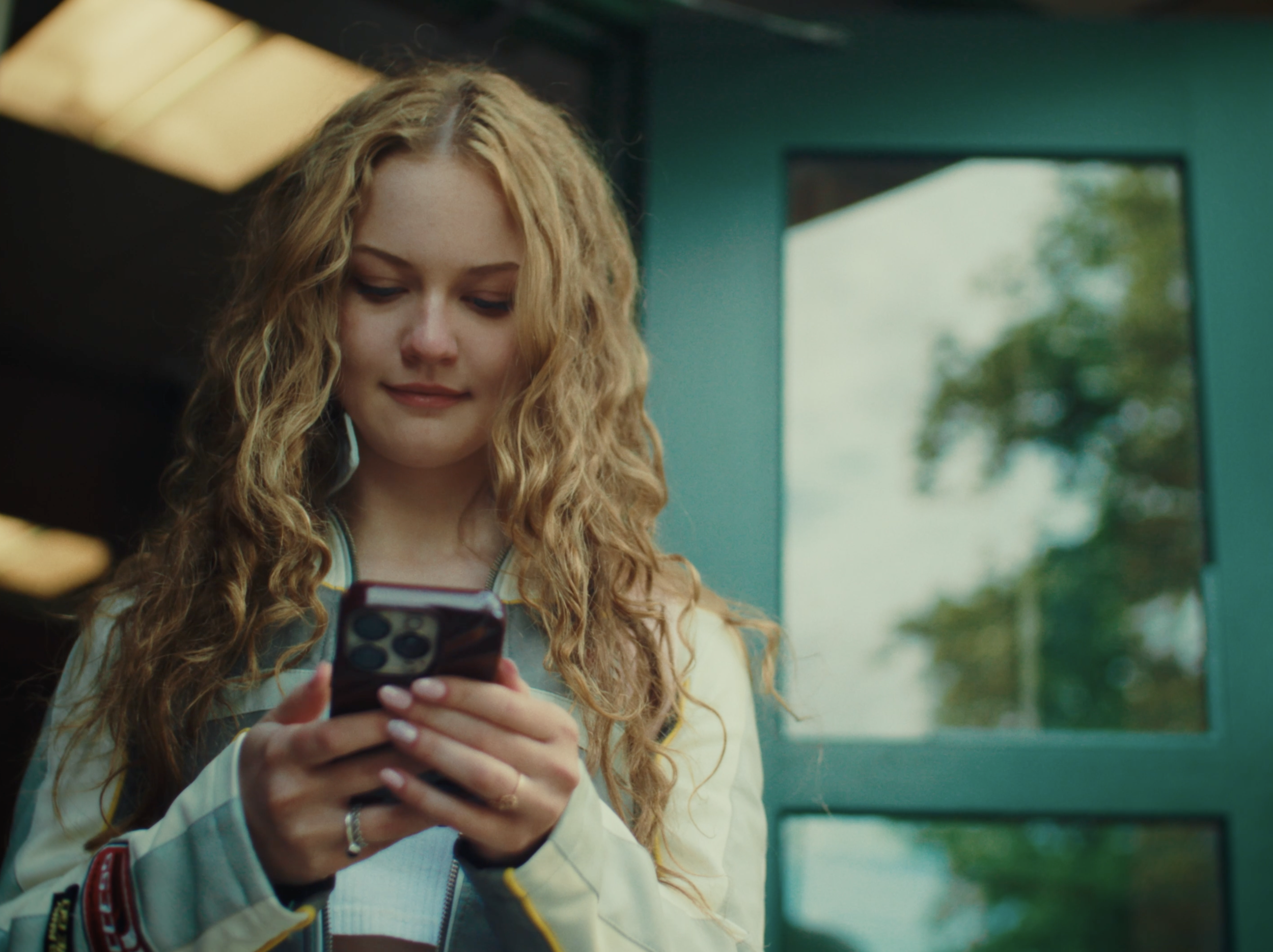 A young woman with long, curly blonde hair smiling while looking at her smartphone outdoors near a teal-colored building.