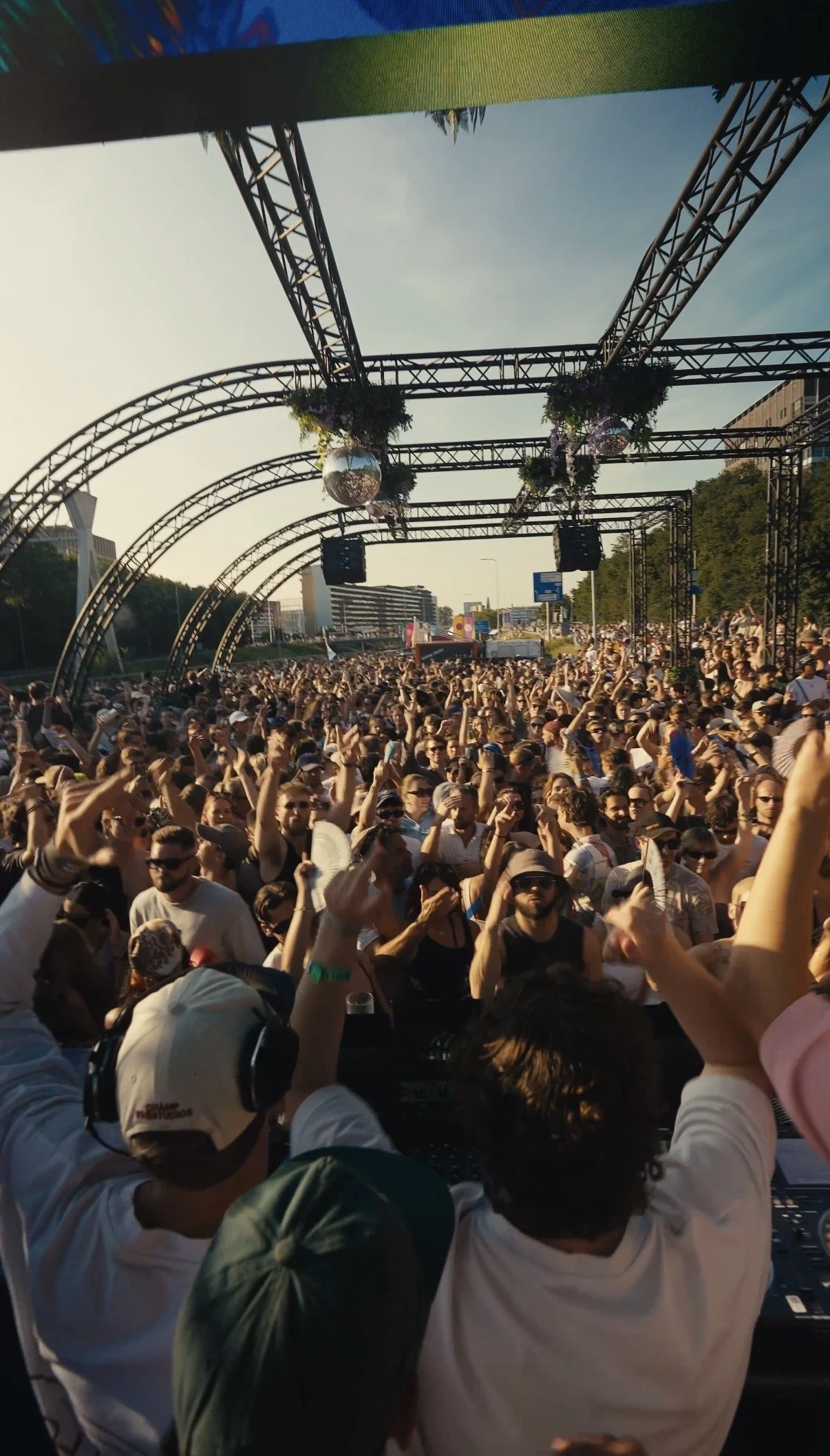 Crowd of people dancing and enjoying music at an outdoor music festival stage with metal arches and hanging disco balls, during sunset.