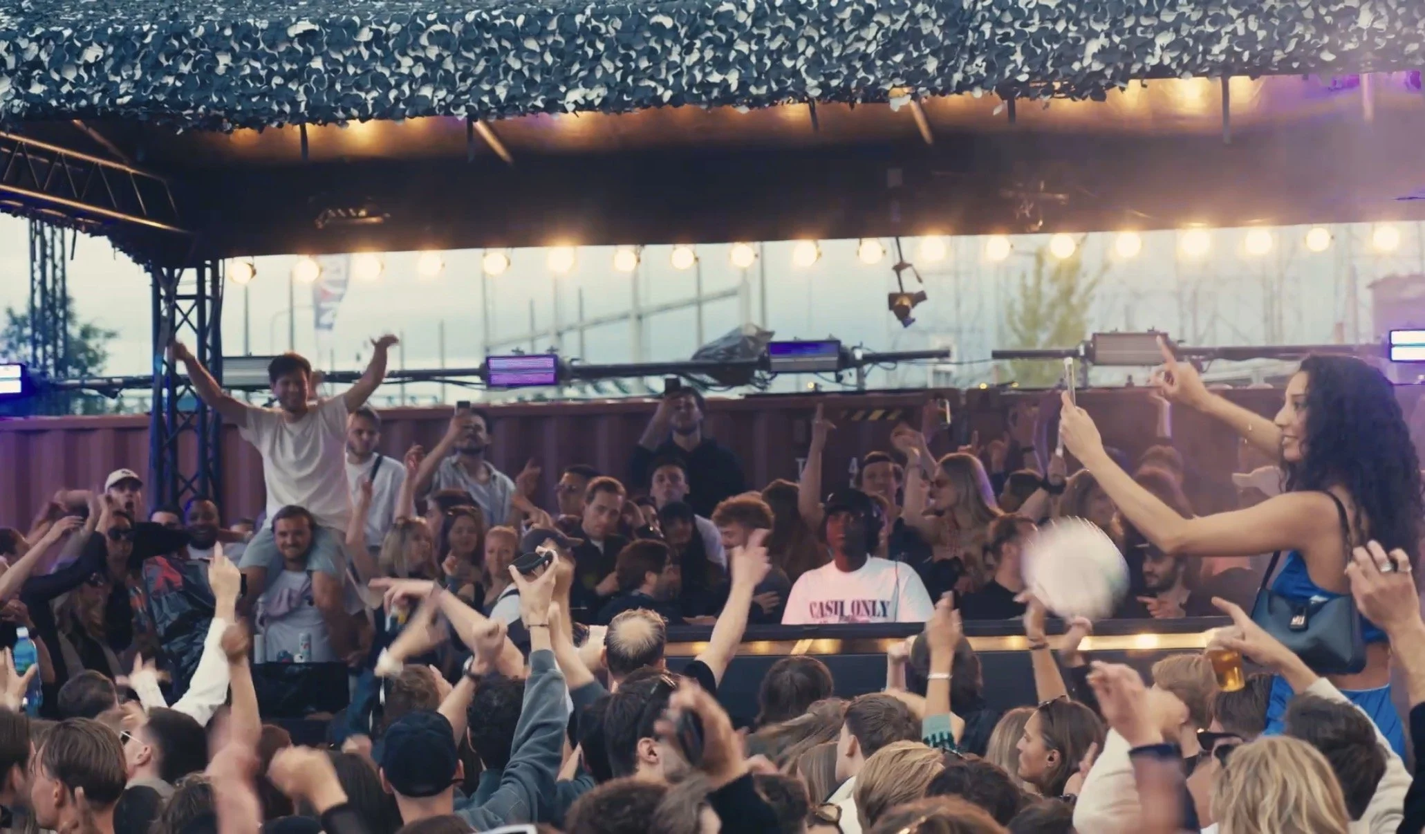 Crowd of people at a music concert or festival, with some dancing and taking photos, under a canopy with hanging string lights.