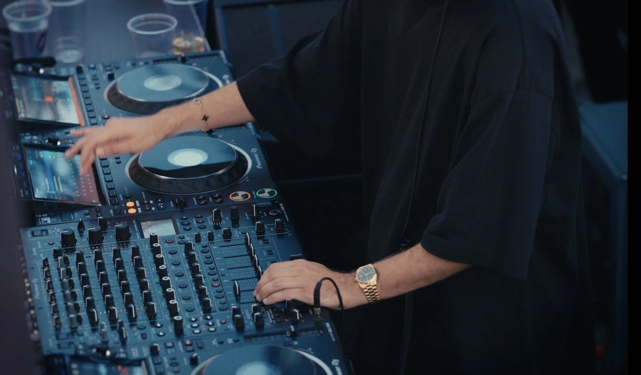 A DJ wearing a black shirt and a gold watch, operating a professional DJ mixer and turntables at a club or event.