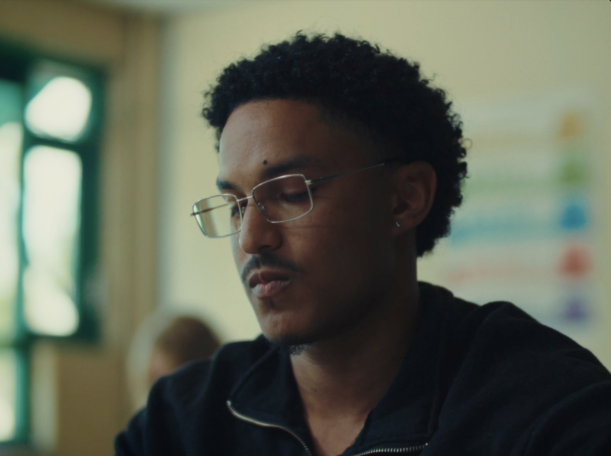 A young man with curly black hair, glasses, and earrings looks down thoughtfully in a classroom setting with a colorful chart on the wall behind him.