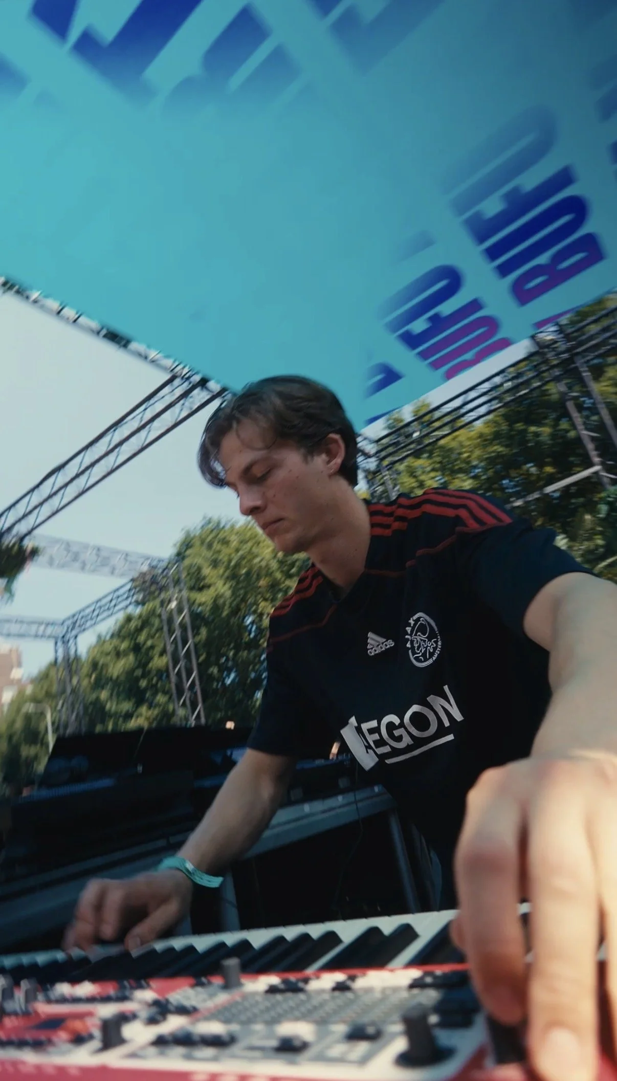 A young male DJ with dark hair, wearing a black jersey with red stripes and a logo, is operating DJ equipment outdoors at a festival. There are large stage banners and trees in the background, under a clear sky.