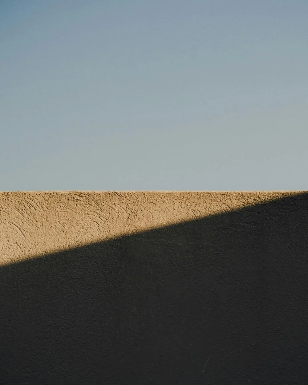 Close-up of a textured beige wall with a shadow cast on it, under a clear blue sky.