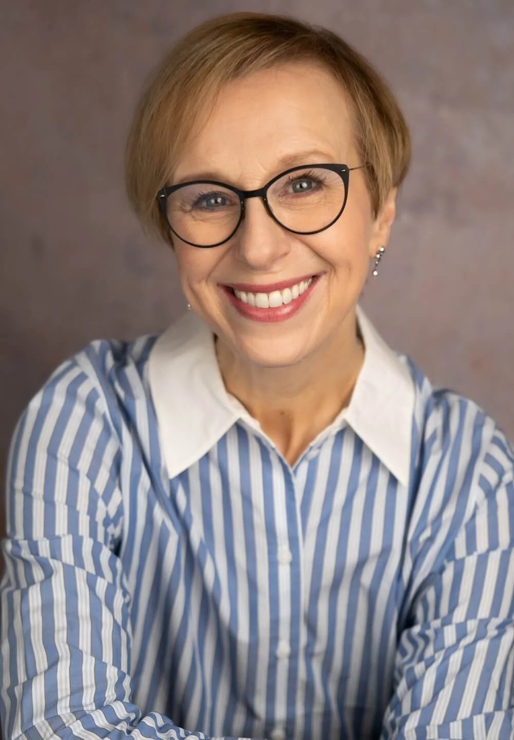 A smiling woman with short light brown hair, wearing black glasses, a white collared shirt, and a blue and white striped blazer, posing against a neutral background.
