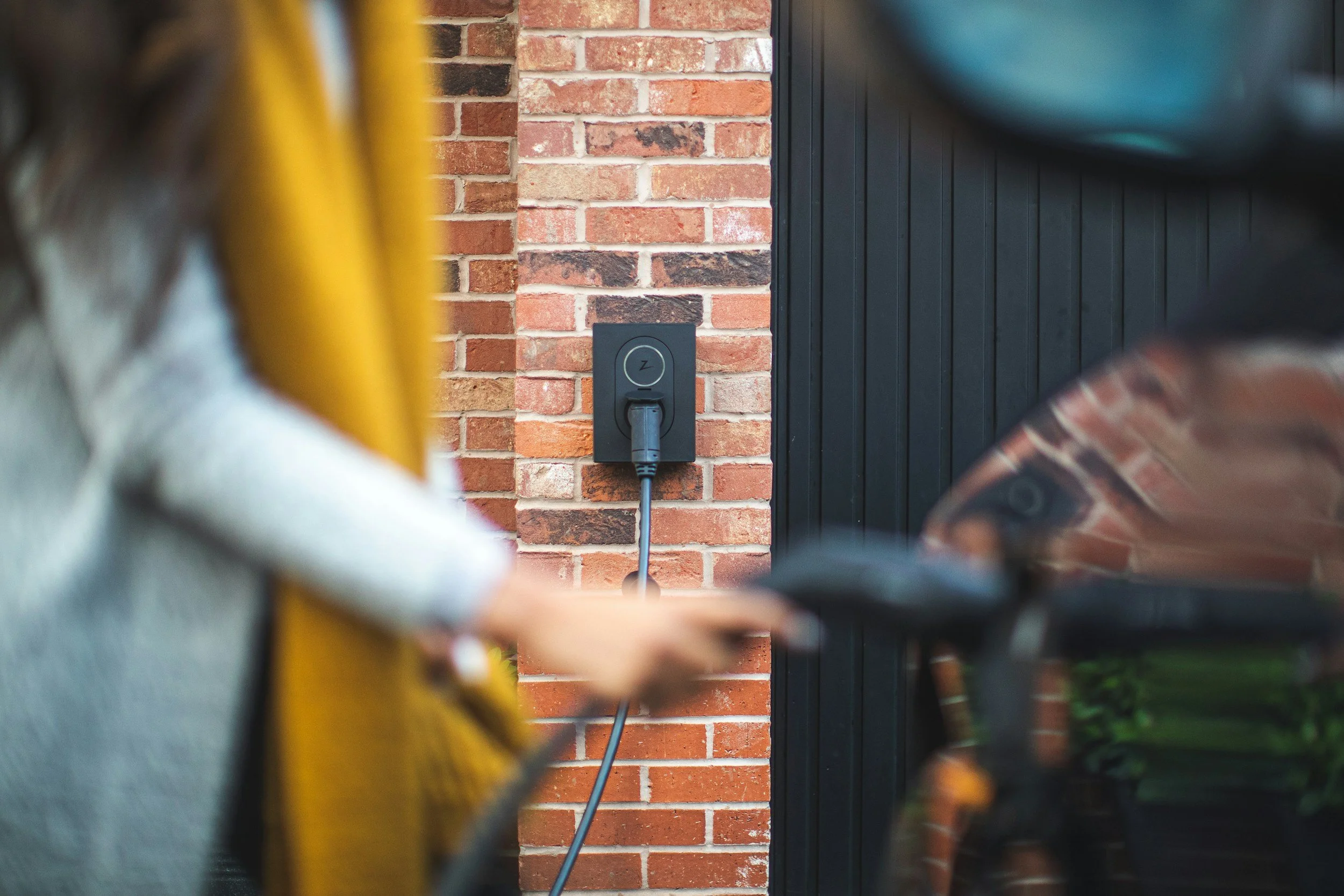 Close-up of an electric vehicle charging station mounted on a brick wall, with a charging cable plugged in, and two people with blurred faces in the foreground, one on a bicycle and the other holding a mobile phone.