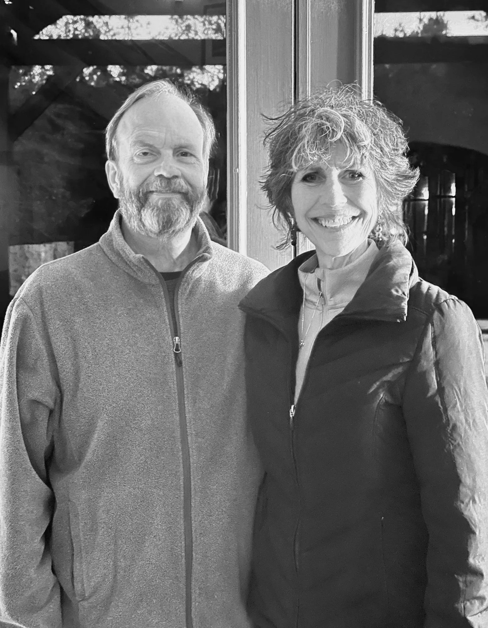 A man and a woman standing indoors, both smiling and looking at the camera. The man has a beard and is wearing a zip-up jacket. The woman has curly hair and is wearing a vest over a shirt.