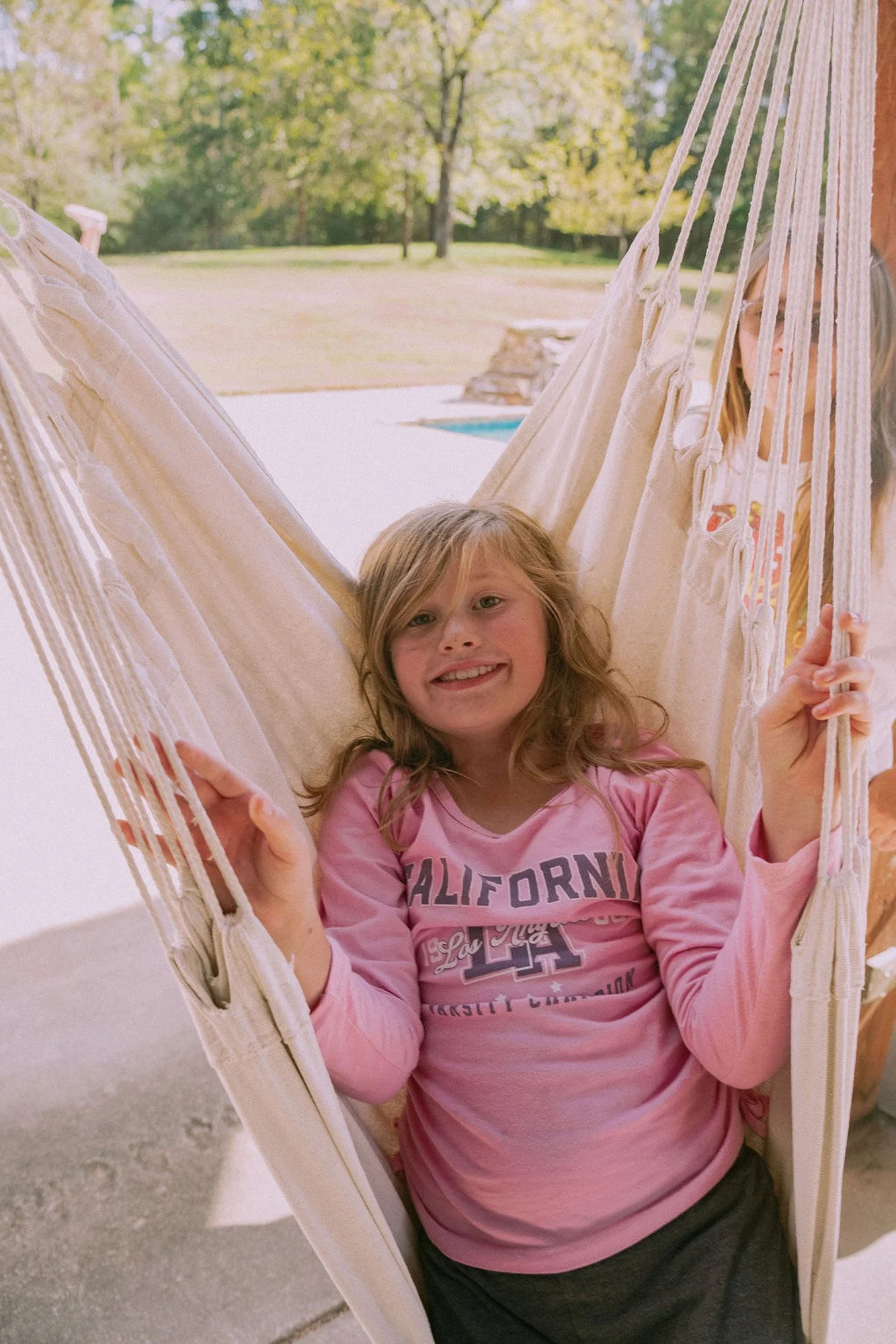 Young girl smiling and relaxing in a hammock outdoors with trees in the background.