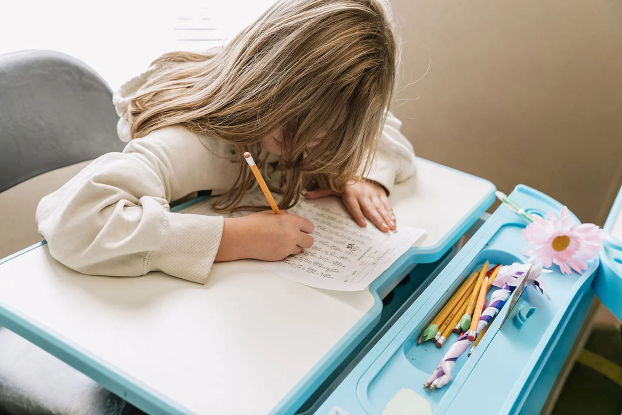 A young girl with long blonde hair writing on a sheet of paper at a school desk. The desk has a built-in tray with pencils, writing utensils, and a pink flower, and she is using a yellow pencil.