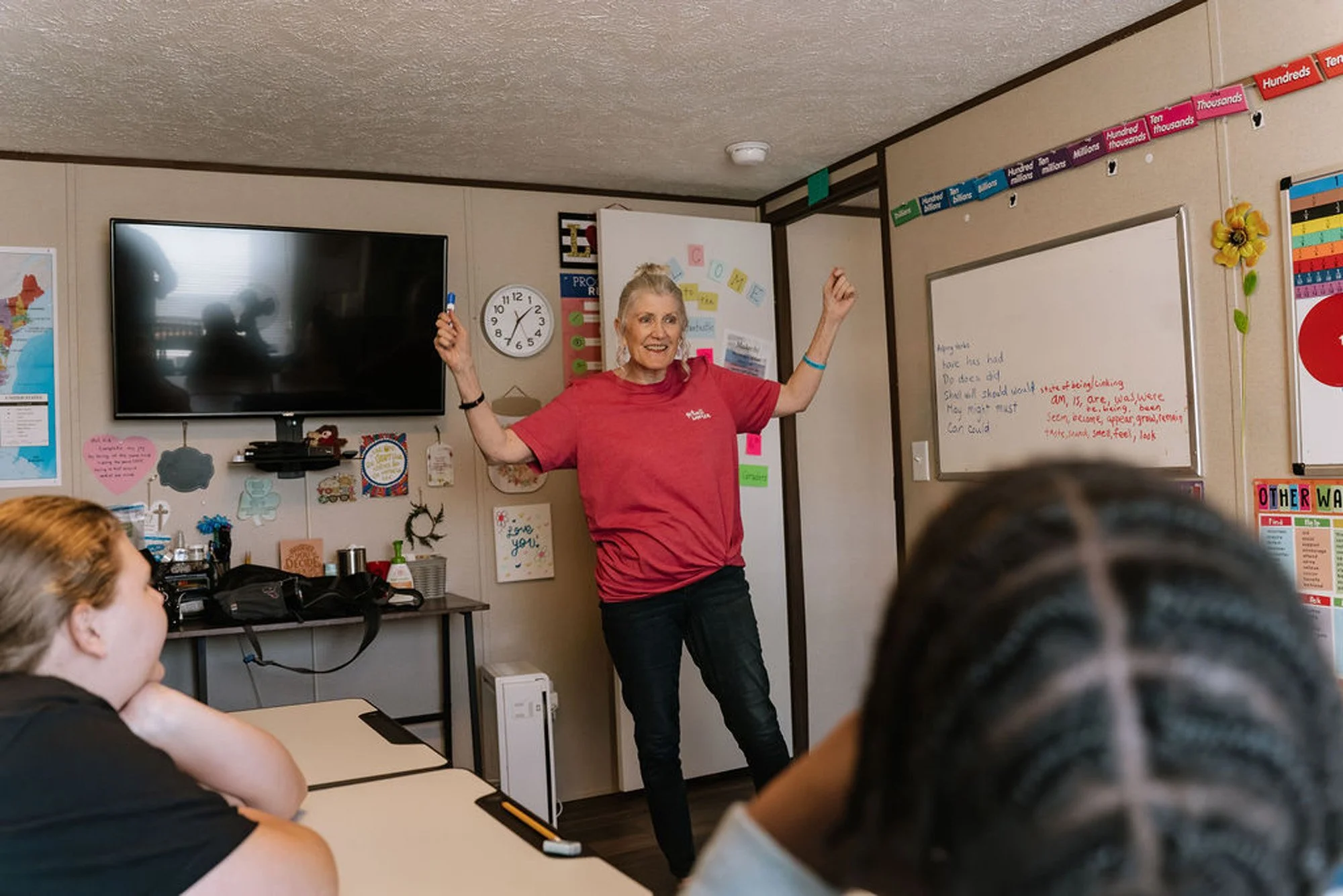A classroom with a teacher standing at the front, gesturing with both arms raised, while students sit at desks listening. The teacher is smiling and wearing a red shirt. The classroom has a whiteboard, a large TV screen, educational posters, and colorful banners.