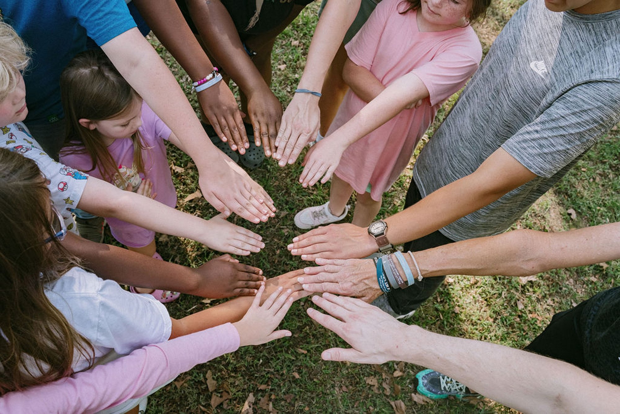 Group of children and adults with hands extended inward, palms touching or nearly touching, outdoors on grass.