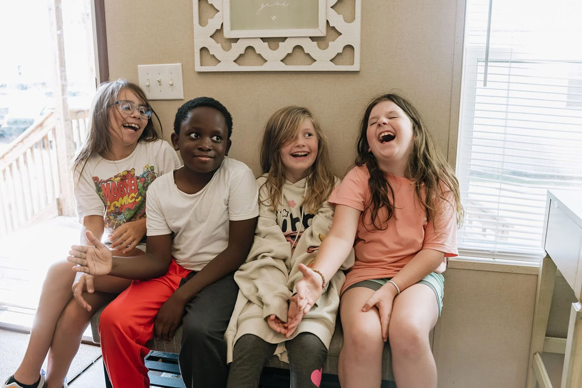 Four children sitting on a bench inside a house, laughing and smiling, with blinds covering a window on the right.