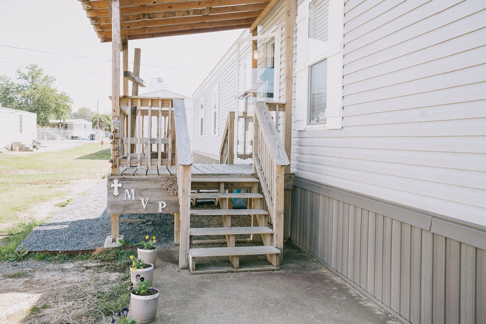 Wooden front porch with stairs and railing attached to a beige house, with potted plants and decorative signs