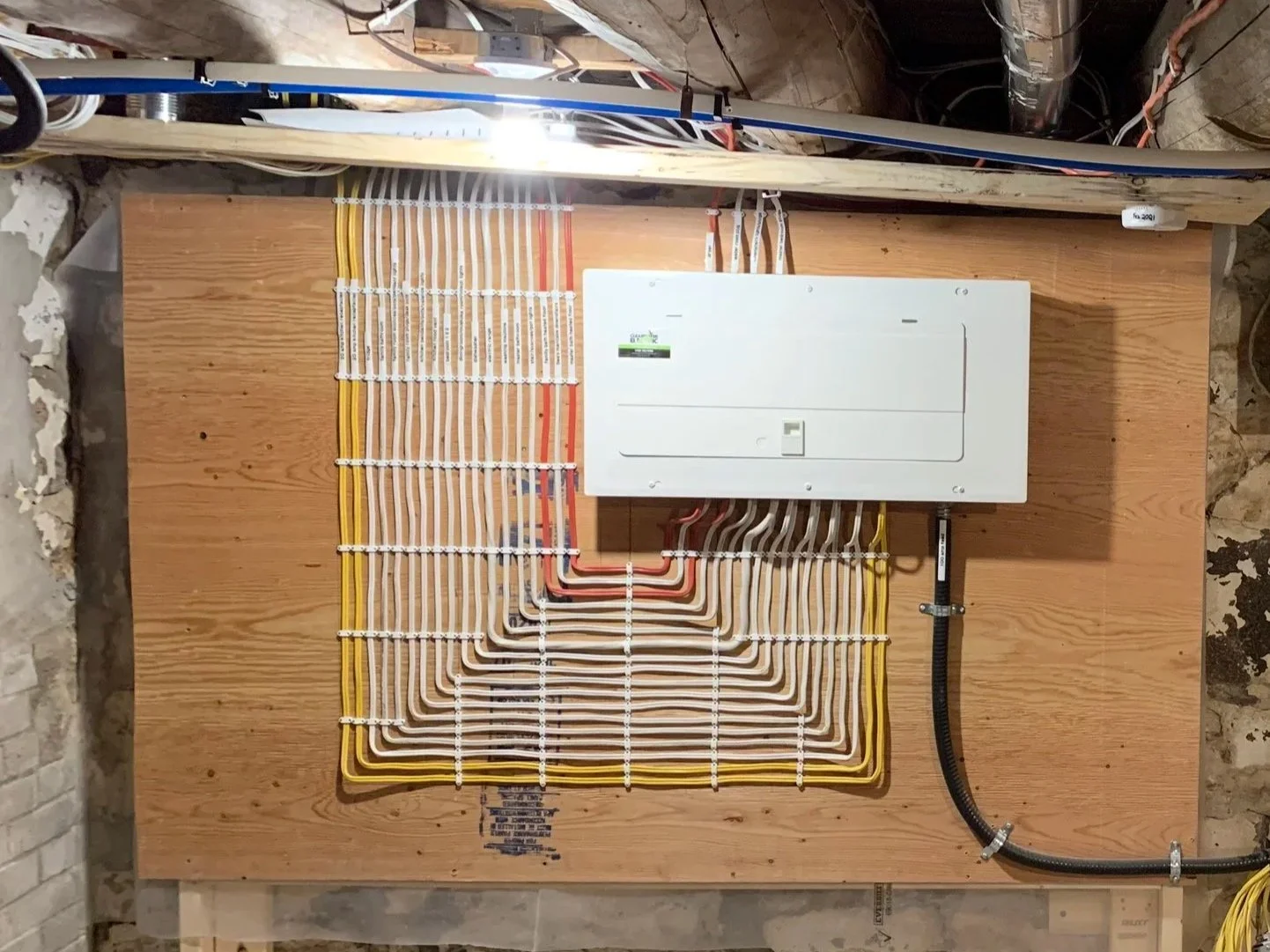 Electrical installation by Clearwater Electric. Electrical panel and wiring setup on a wooden board behind a brick wall with exposed pipes and concrete floor.